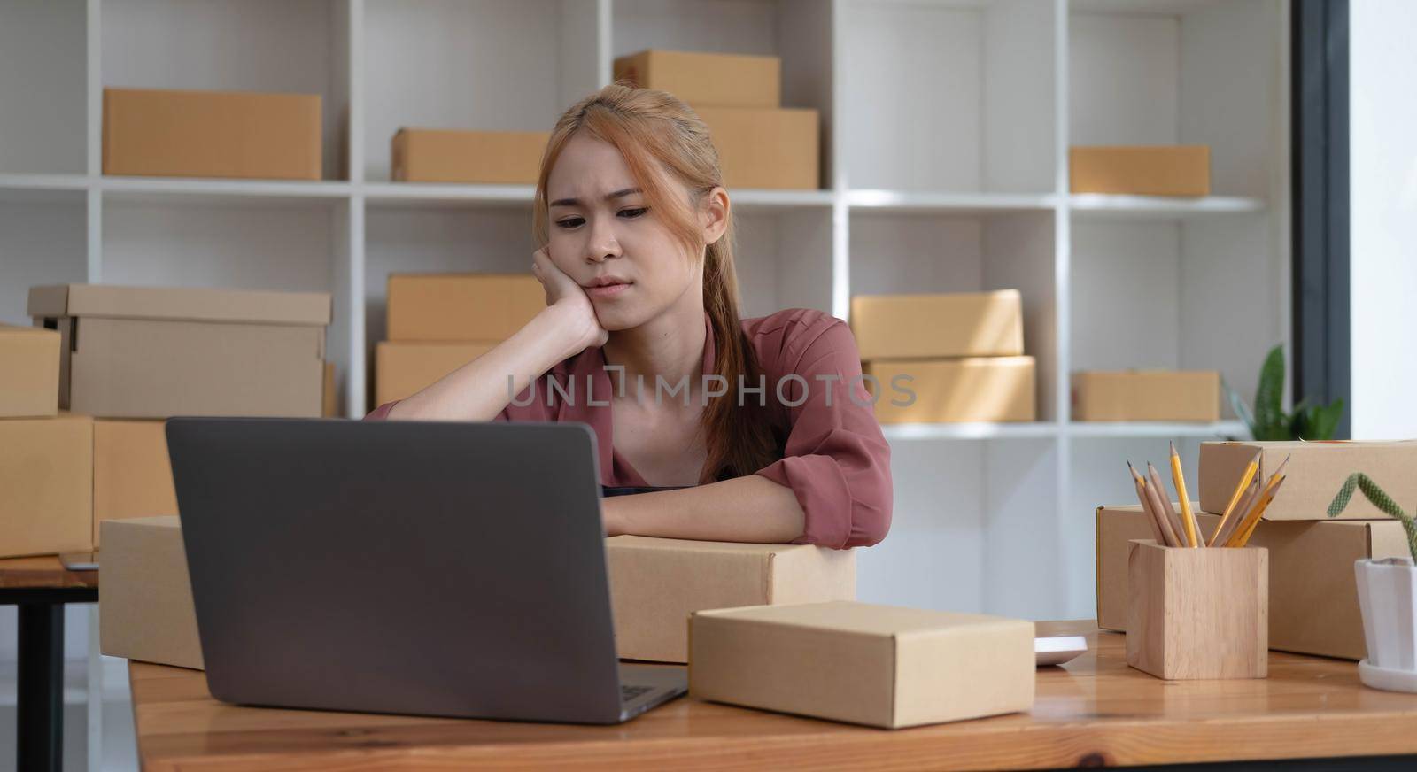 A portrait of young serious Asian woman working with laptop in the office full of packages and boxes stacking up, busy looking table, for SME, delivery, start up business and home office concept.