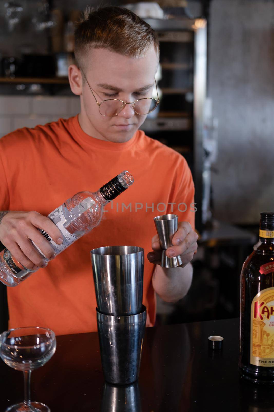 stylish young man hipster in orange t-shirt making mixing a cocktail in a dark loft cafe. alcohol drink in modern bar by oliavesna