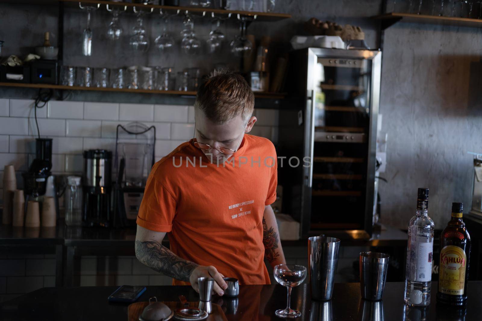 stylish young man hipster in orange t-shirt making mixing a cocktail in a dark loft cafe. alcohol drink in modern bar.
