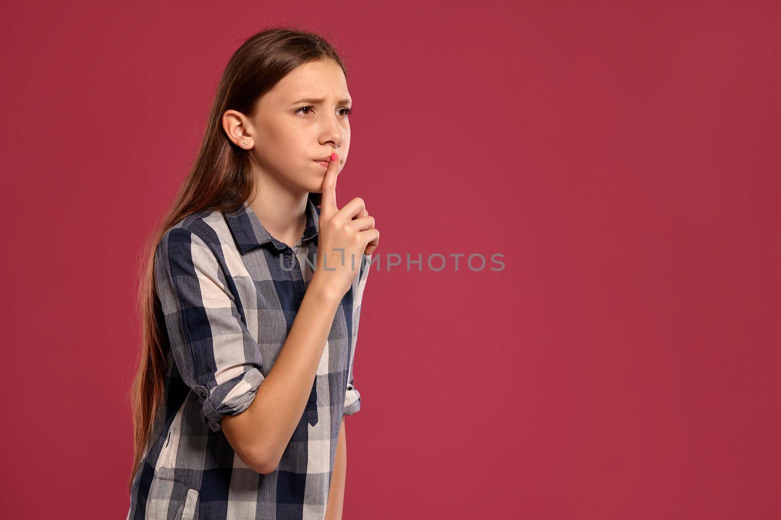Portrait of a gorgeous teenage maiden in a casual checkered shirt asking someone to be quiet while posing against a pink studio background. Long hair, healthy clean skin and brown eyes. Sincere emotions concept. Copy space.
