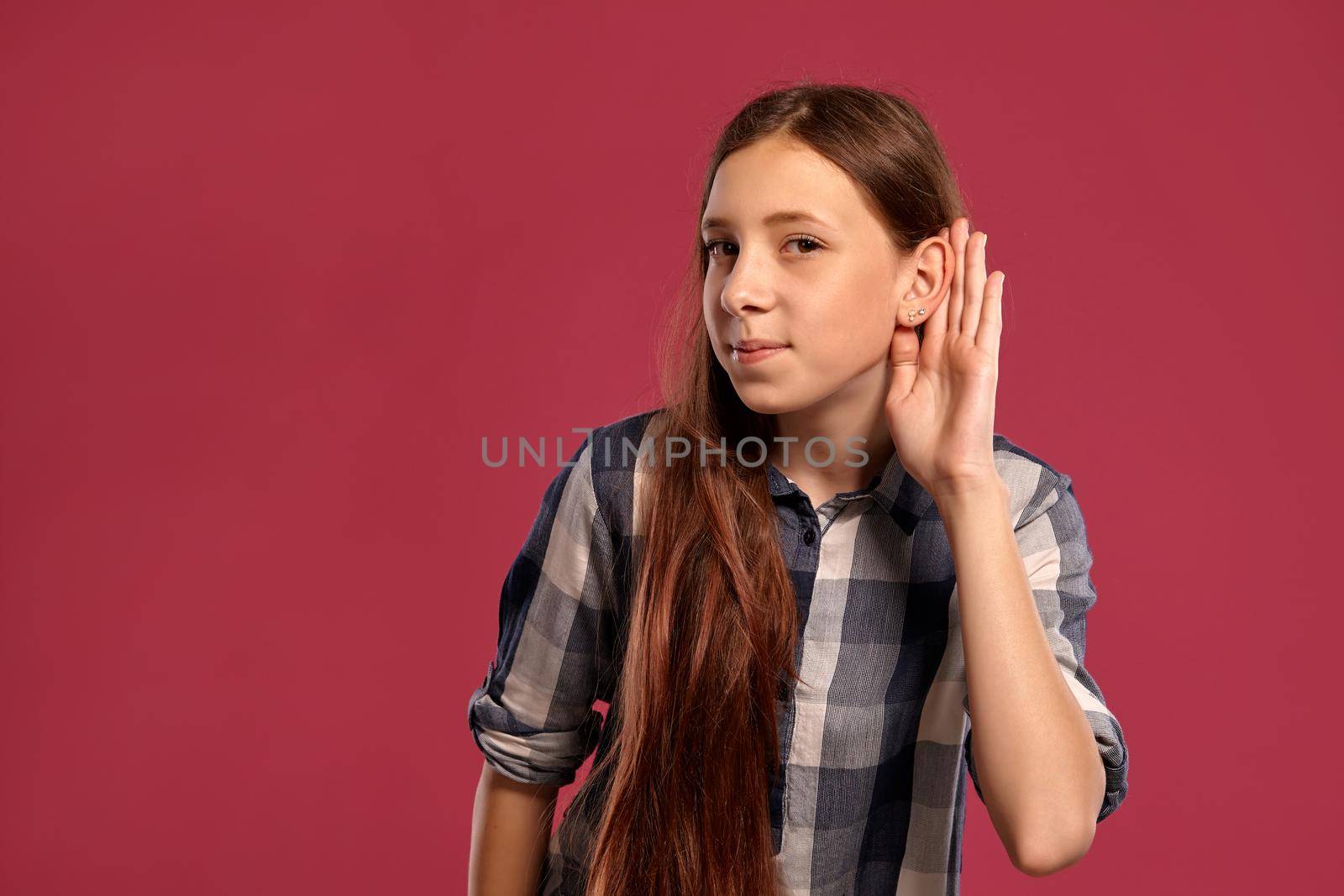 Portrait of a lovely teenager in a casual checkered shirt listening someone while posing against a pink studio background. Long hair, healthy clean skin and brown eyes. Sincere emotions concept. Copy space.