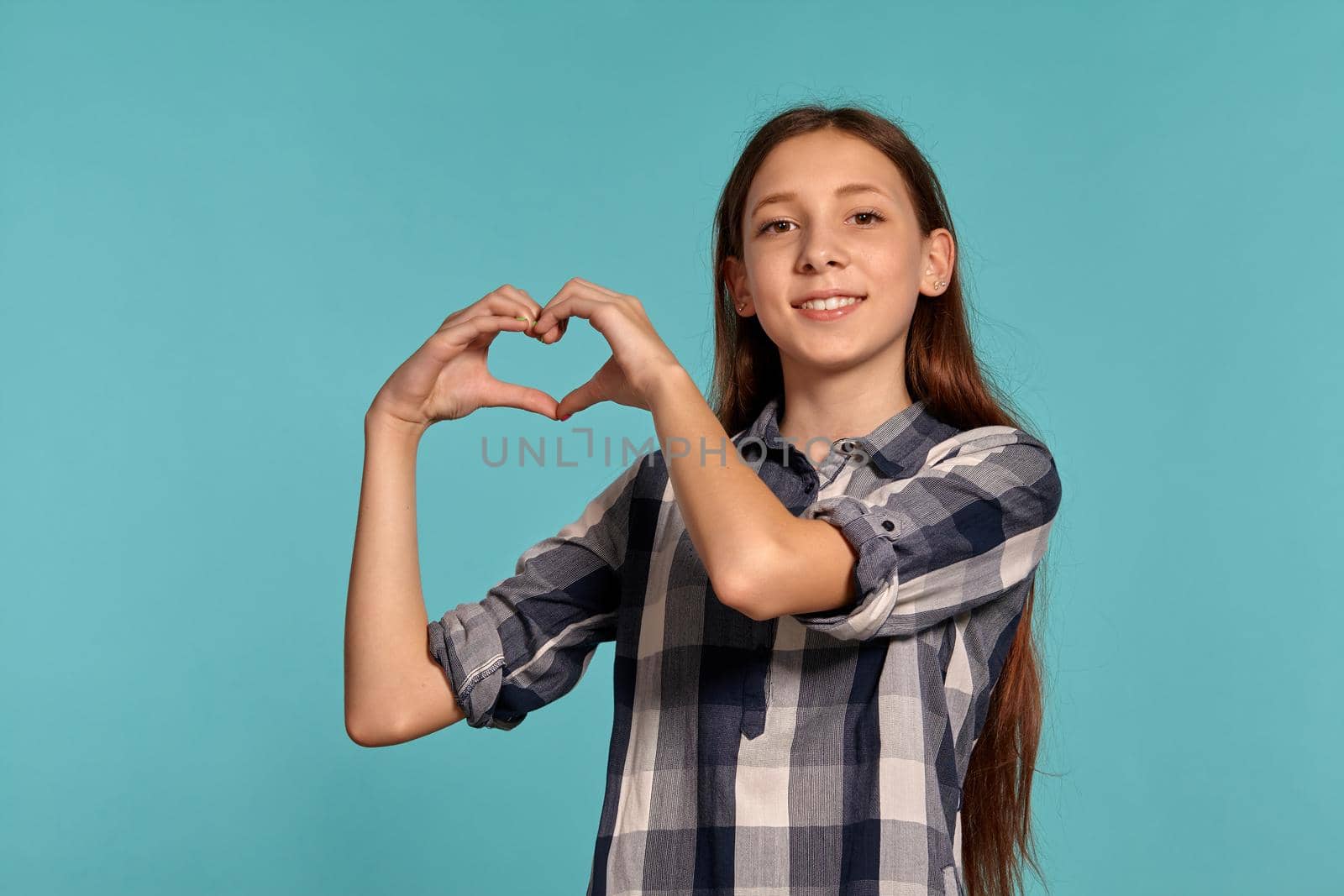 Portrait of a gorgeous teenage girl in a casual checkered shirt showing a heart sign of her folded fingers and posing against a blue studio background. Long hair, healthy clean skin and brown eyes. Sincere emotions concept. Copy space.