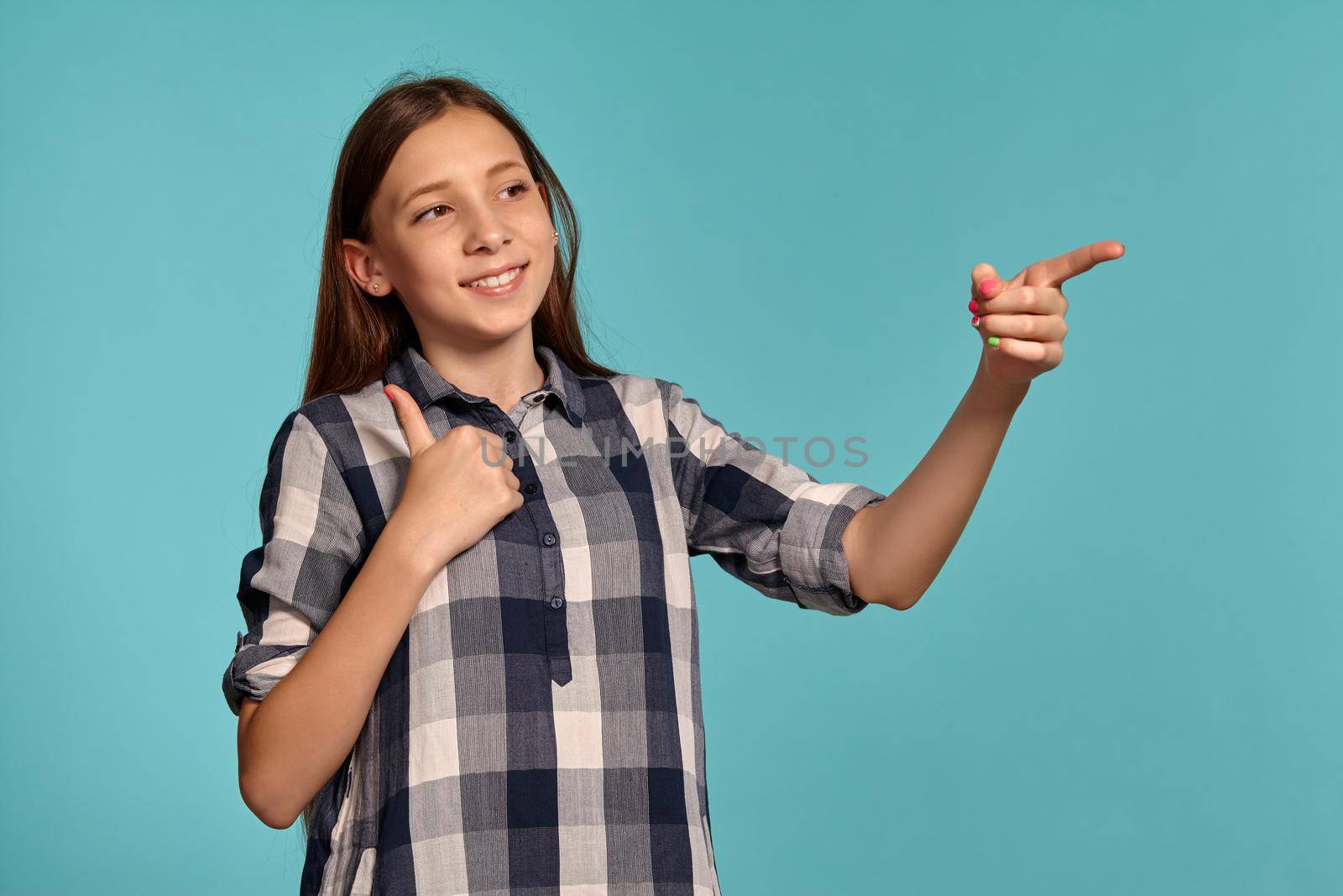 Portrait of a charming teenage lady in a casual checkered shirt pointing at something while posing against a blue studio background. Long hair, healthy clean skin and brown eyes. Sincere emotions concept. Copy space.