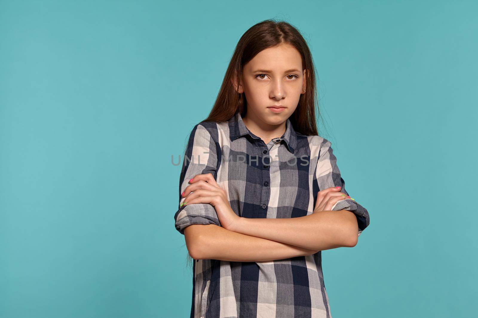 Portrait of a charming teenage girl in a casual checkered shirt embraced herself and looking upset while posing against a blue studio background. Long hair, healthy clean skin and brown eyes. Sincere emotions concept. Copy space.