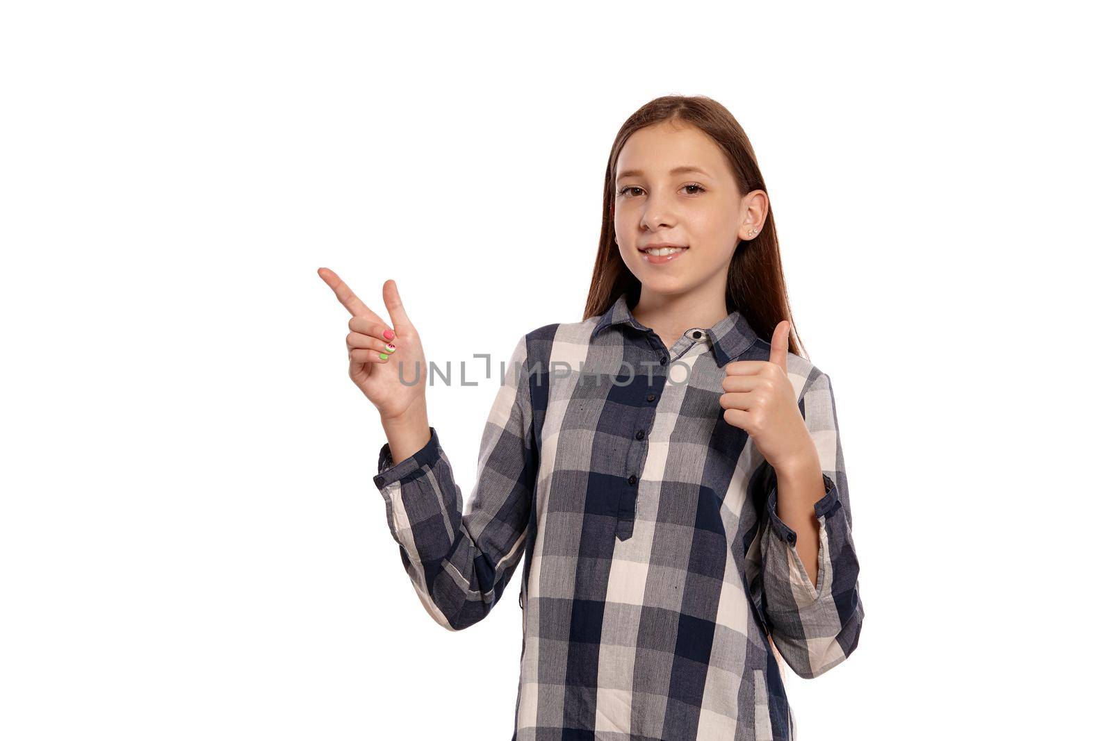 Portrait of a cute teenage female in a casual checkered shirt pointing at something and raised thumb up whie posing isolated on white studio background. Long hair, healthy clean skin and brown eyes. Sincere emotions concept. Copy space.
