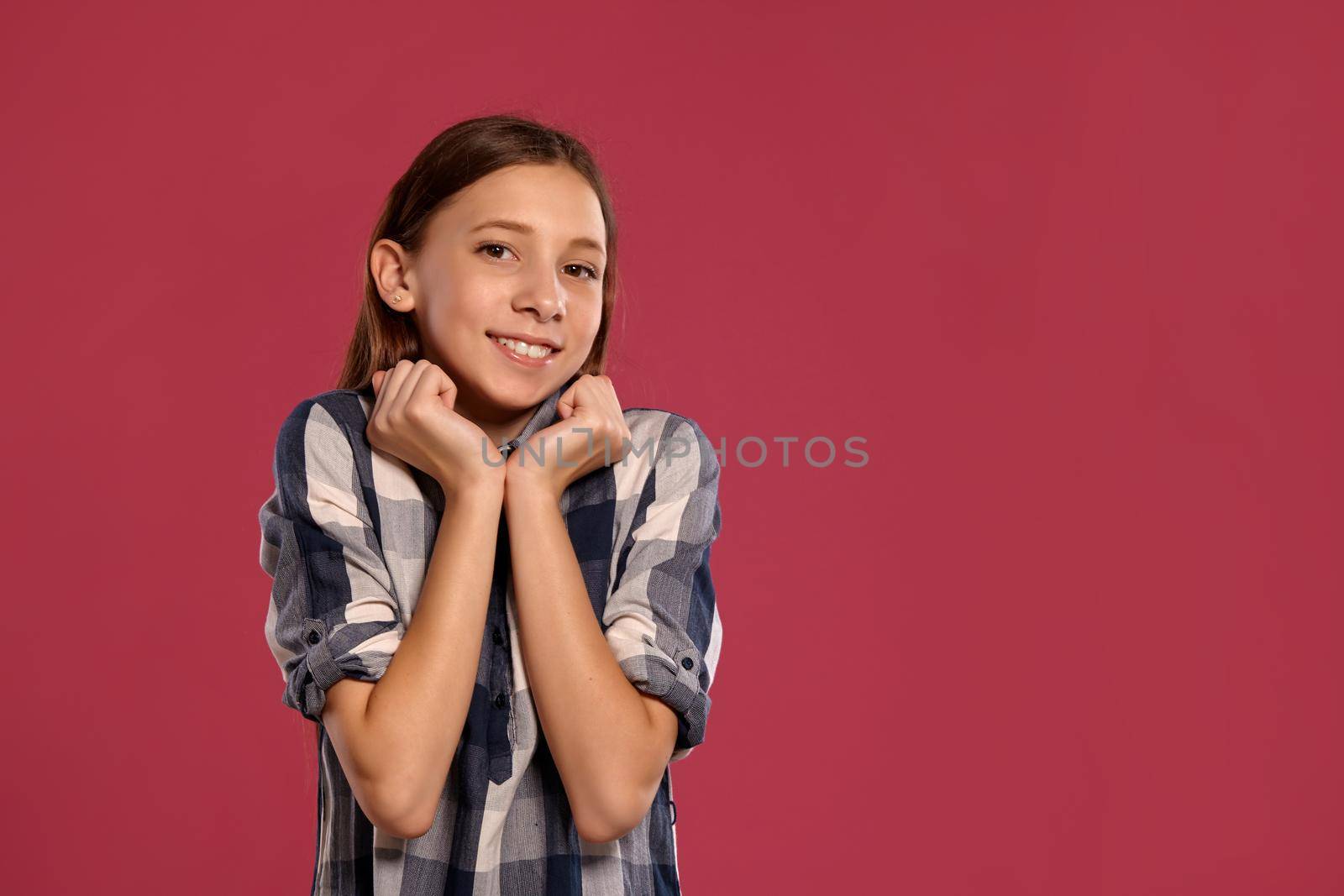 Portrait of a beautiful teenage girl in a casual checkered shirt smiling and looking at the camera while posing against a pink studio background. Long hair, healthy clean skin and brown eyes. Sincere emotions concept. Copy space.