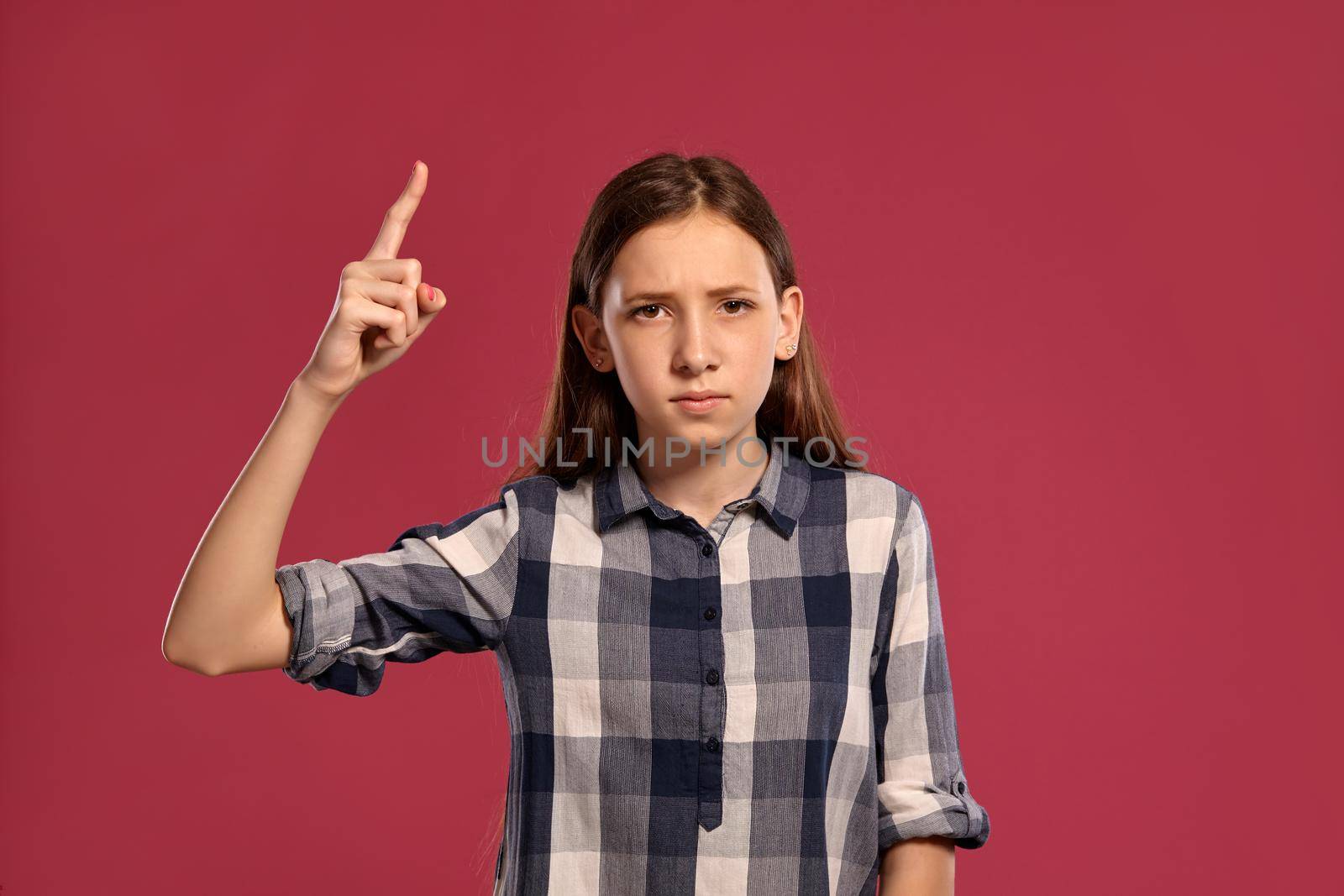 Portrait of a gorgeous teenage lady in a casual checkered shirt raised her index finger up and looking at the camera while posing against a pink studio background. Long hair, healthy clean skin and brown eyes. Sincere emotions concept. Copy space.