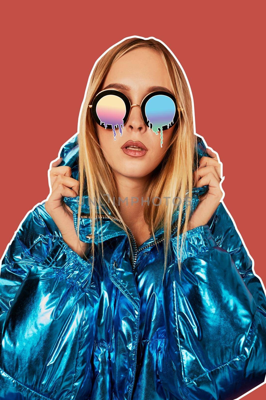 Close-up fashion portrait of an alluring girl in a warm shiny jacket and cartoon melting glasses, holding her hood and posing against a pink background of studio. Art collage.