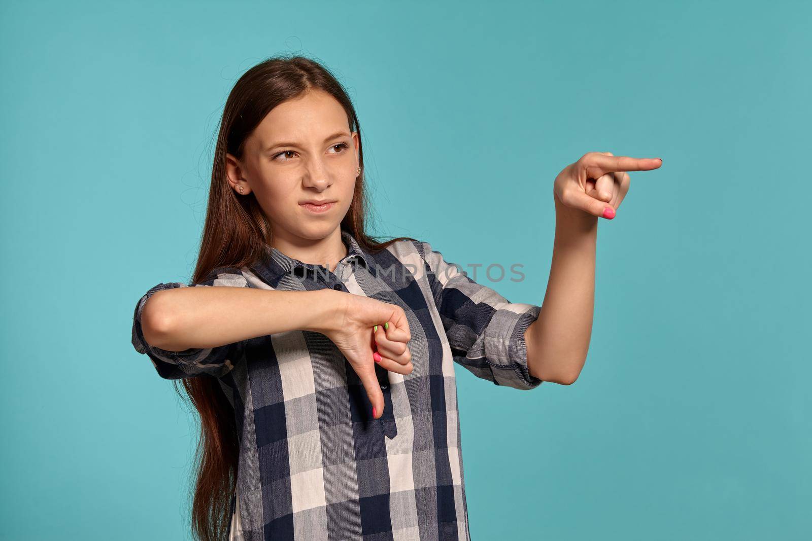 Portrait of a charming teenage female in a casual checkered shirt pointing at someone and looking away while posing against a blue studio background. Long hair, healthy clean skin and brown eyes. Sincere emotions concept. Copy space.