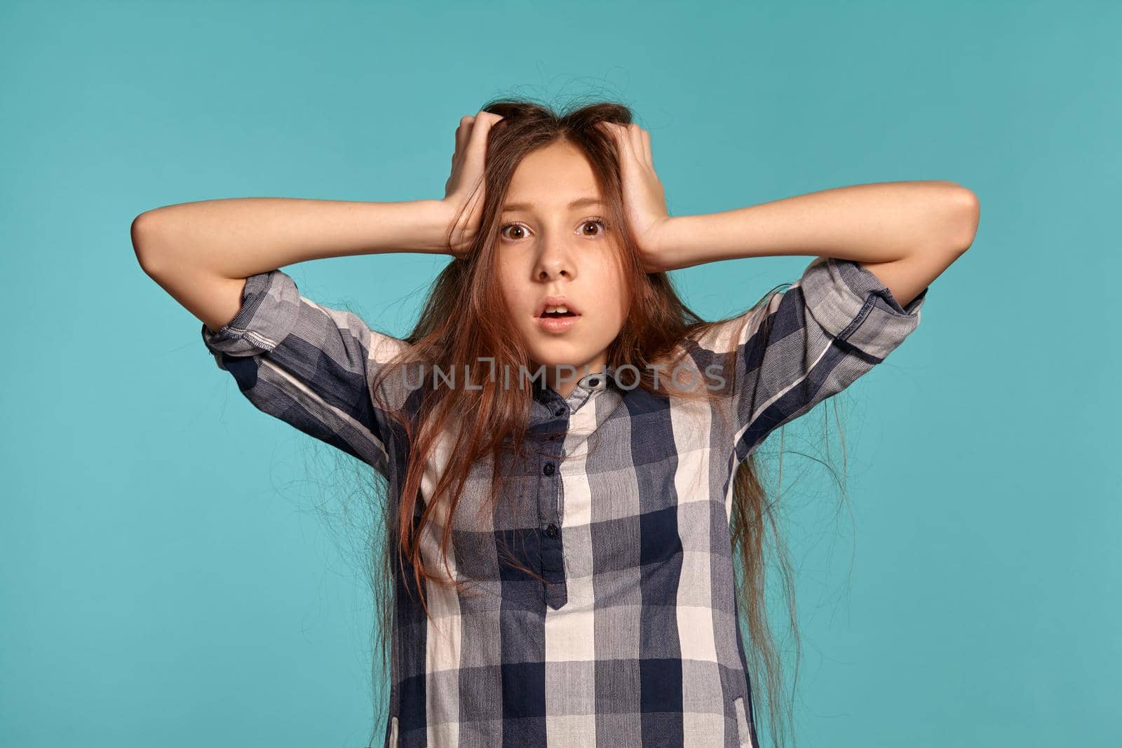 Portrait of a beautiful teenage girl in a casual checkered shirt holding her head and posing against a blue studio background. Long hair, healthy clean skin and brown eyes. Sincere emotions concept. Copy space.