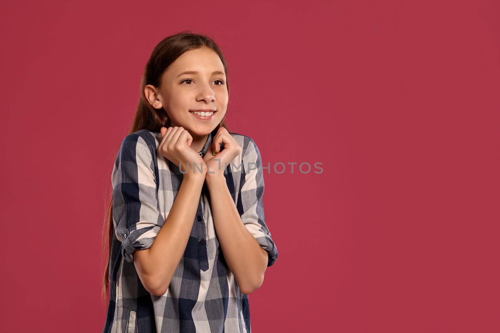 Portrait of a cute teenage maiden in a casual checkered shirt looking happy and posing against a pink studio background. Long hair, healthy clean skin and brown eyes. Sincere emotions concept. Copy space.