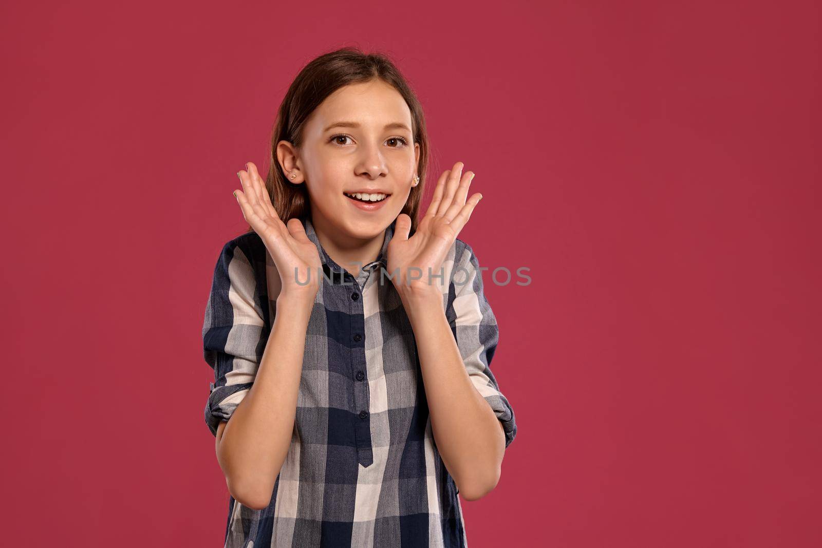 Portrait of a cute teenage female in a casual checkered shirt looking wondered and smiling while posing against a pink studio background. Long hair, healthy clean skin and brown eyes. Sincere emotions concept. Copy space.