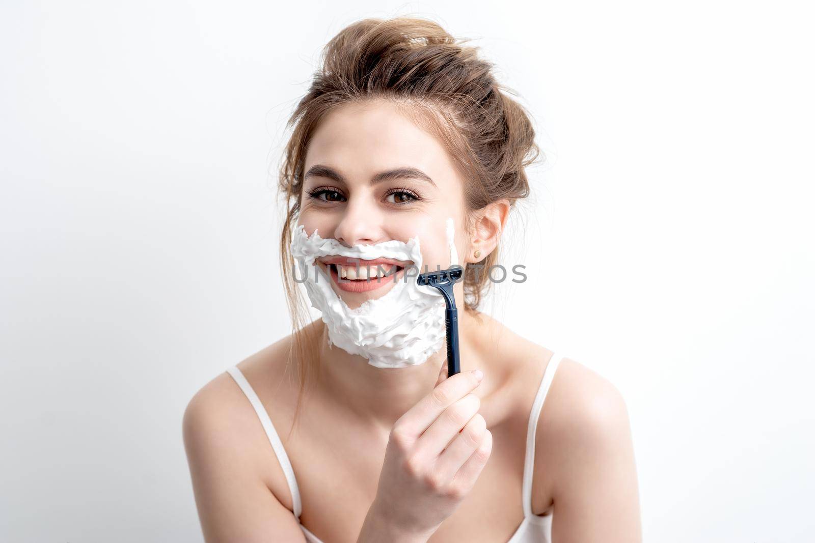 Beautiful young caucasian woman shaving her face by razor on white background. Pretty woman with shaving foam on her face