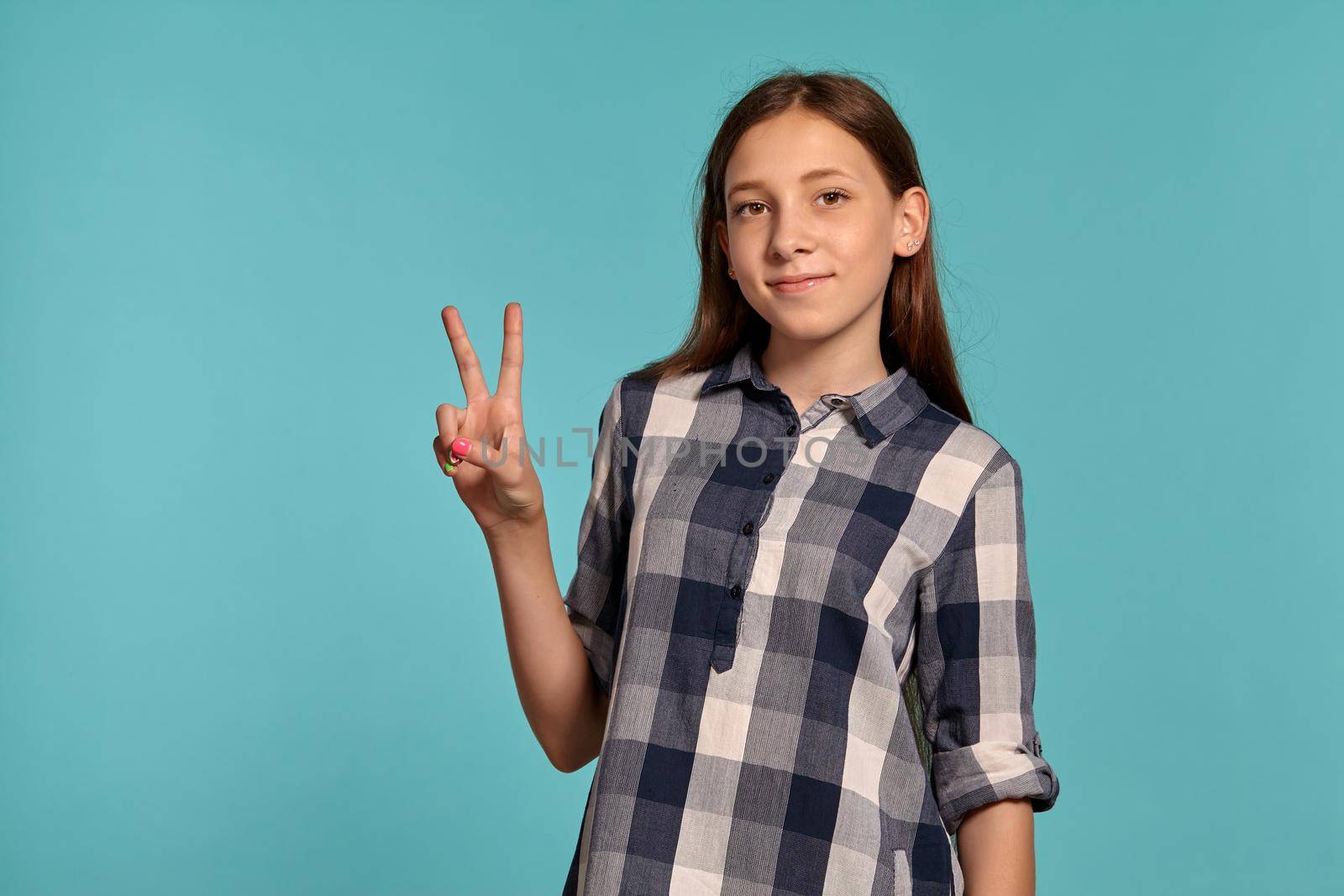Portrait of an attractive teenage girl in a casual checkered shirt showing two fingers and smiling while posing against a blue studio background. Long hair, healthy clean skin and brown eyes. Sincere emotions concept. Copy space.