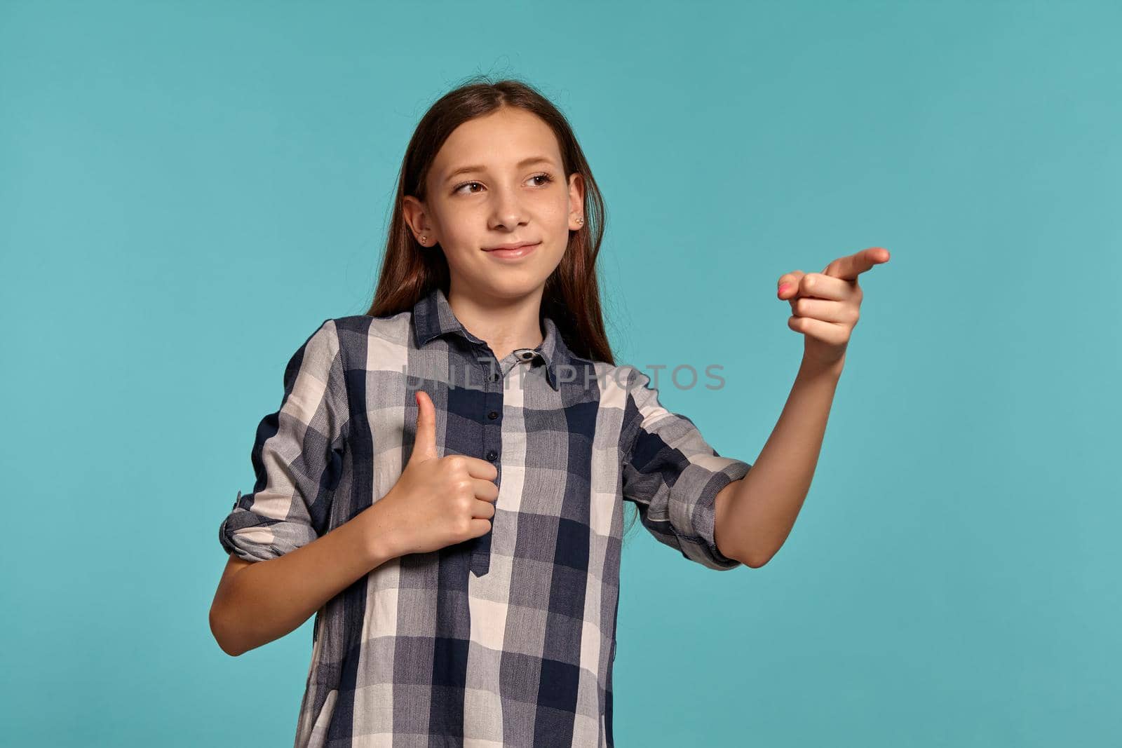 Portrait of a charming teenage maiden in a casual checkered shirt pointing at someone and posing against a blue studio background. Long hair, healthy clean skin and brown eyes. Sincere emotions concept. Copy space.