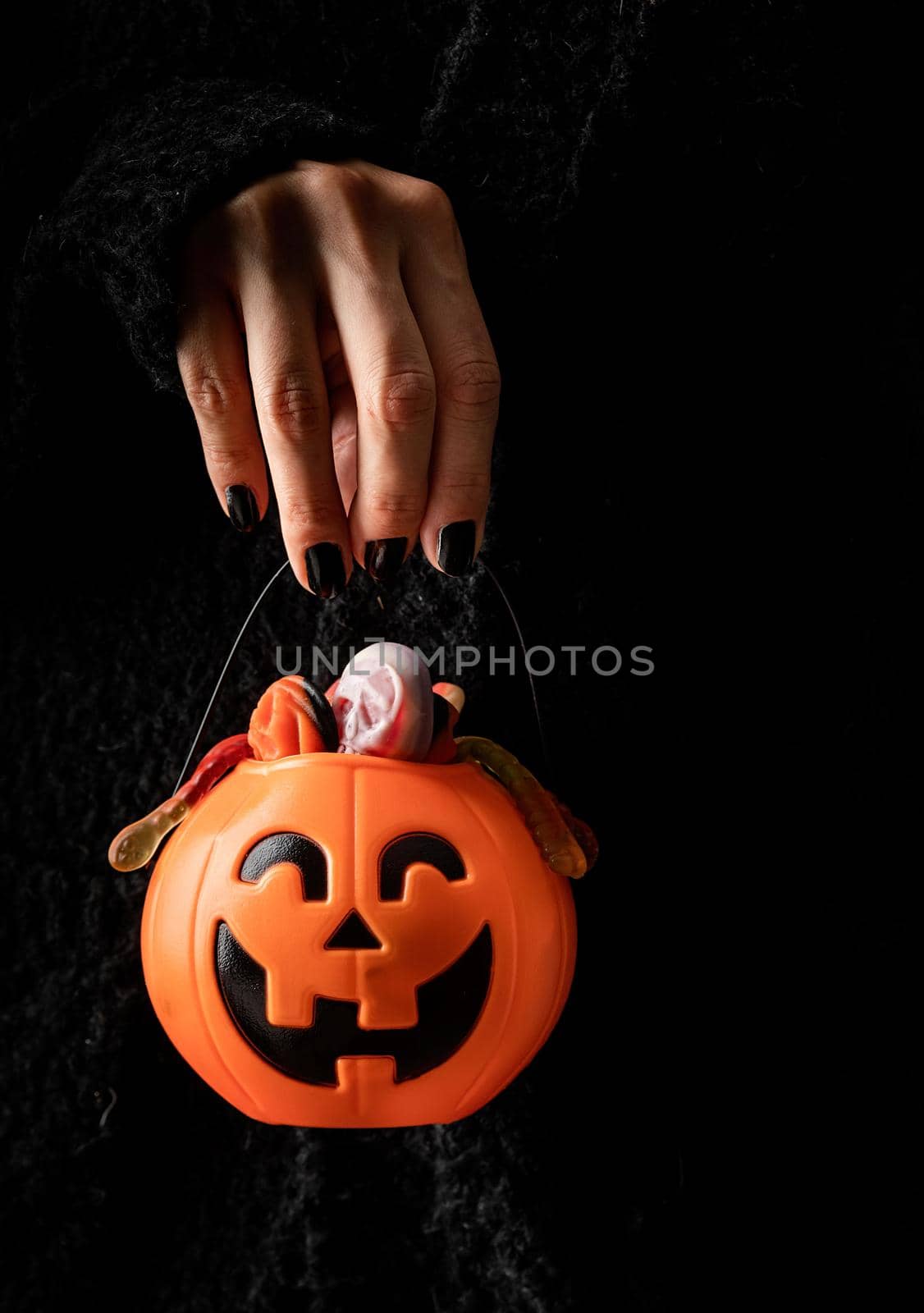 Happy Halloween concept. Scary woman Halloween hand with black nails holding pumpkin full of sweets on dark background with copy space