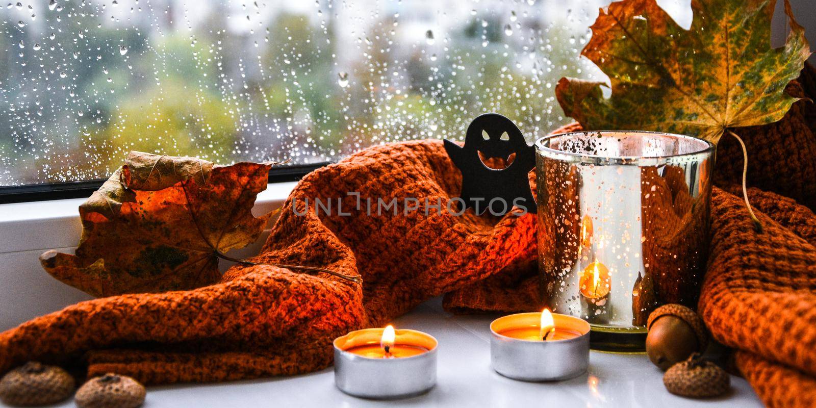 Candles with sweater and ghost pumpkin, dried leaves on windowsill. Halloween home decoration. Rainy window. Cozy home interior