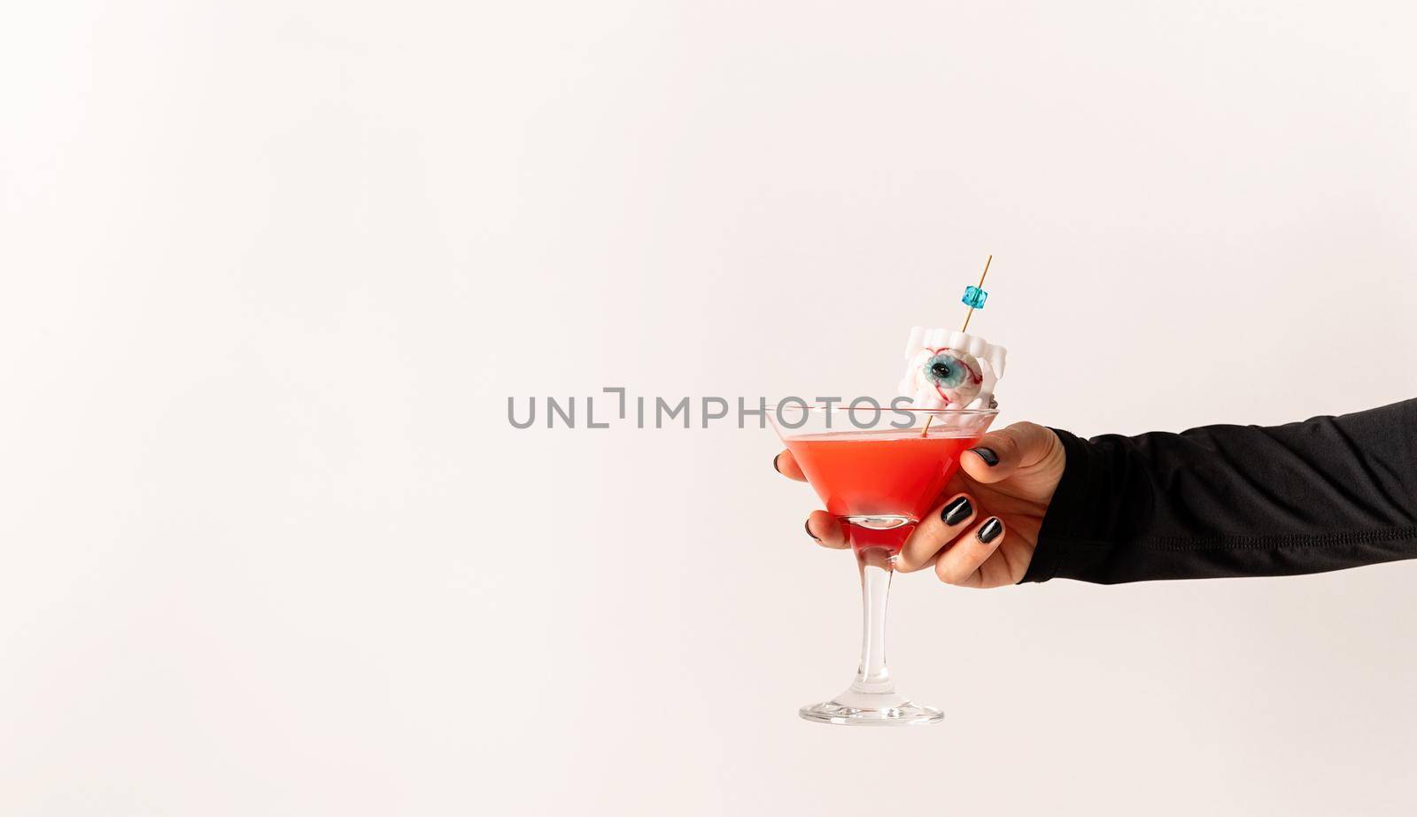 Happy Halloween concept. Woman hand with black nails holding glass with scary red halloween cocktail on white background, copy space