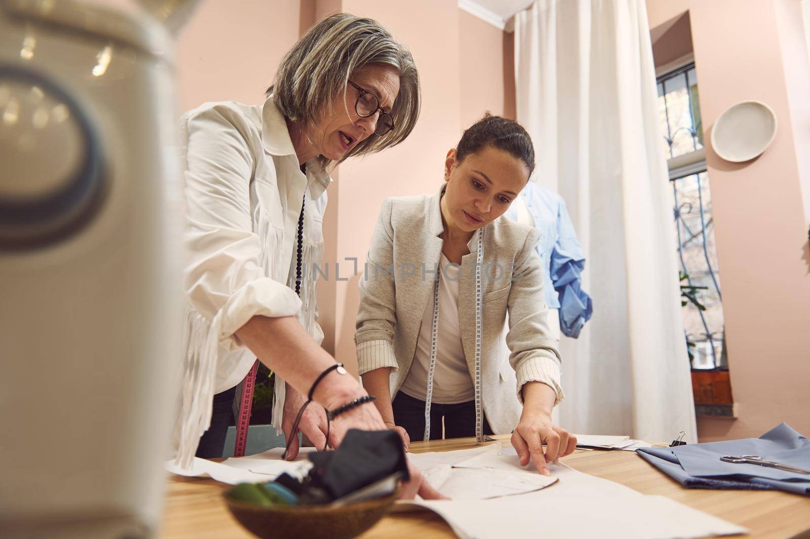 Skilled competent team of two women fashion designers work on collection of sewing patterns to create new collection of stylish modern clothes in a tailoring atelier. Seamstresses discussing patterns