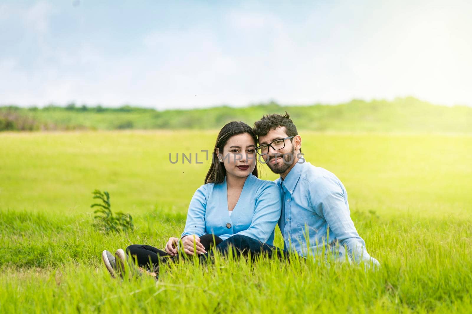 Portrait of a couple in love sitting on the grass in the field, Romantic couple sitting on the grass looking at the camera, Two lovers sitting on the grass looking at the camera by isaiphoto