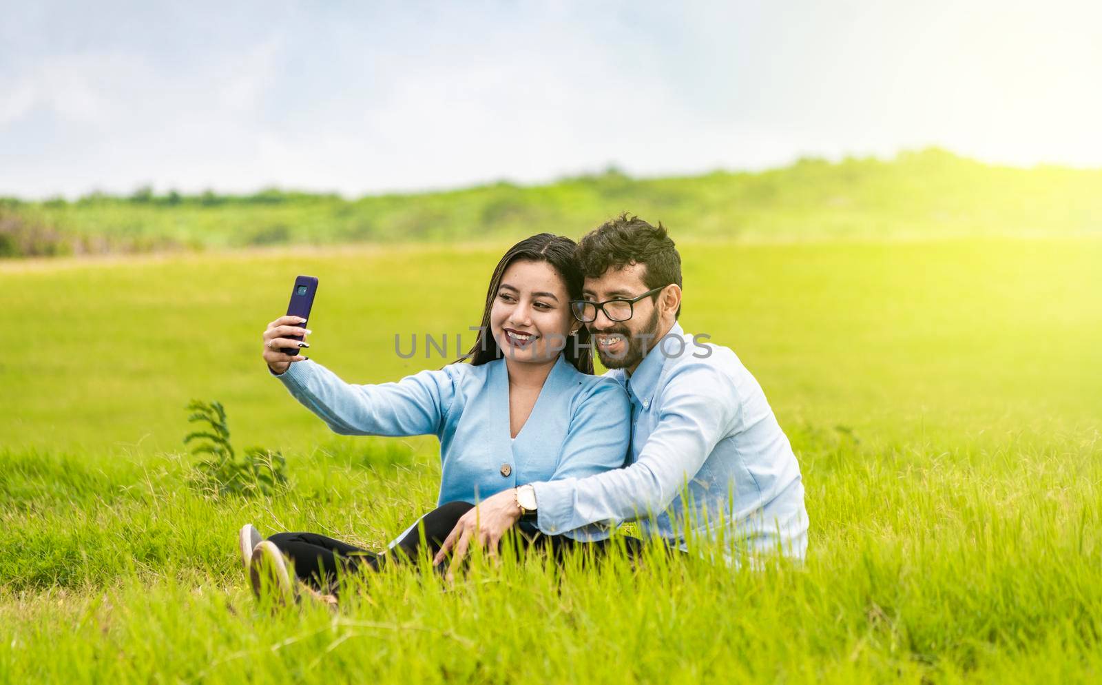 Smiling couple in love sitting on the grass taking selfies, Young couple in love taking a selfie in the field, People in love taking selfies in the field with their smartphone