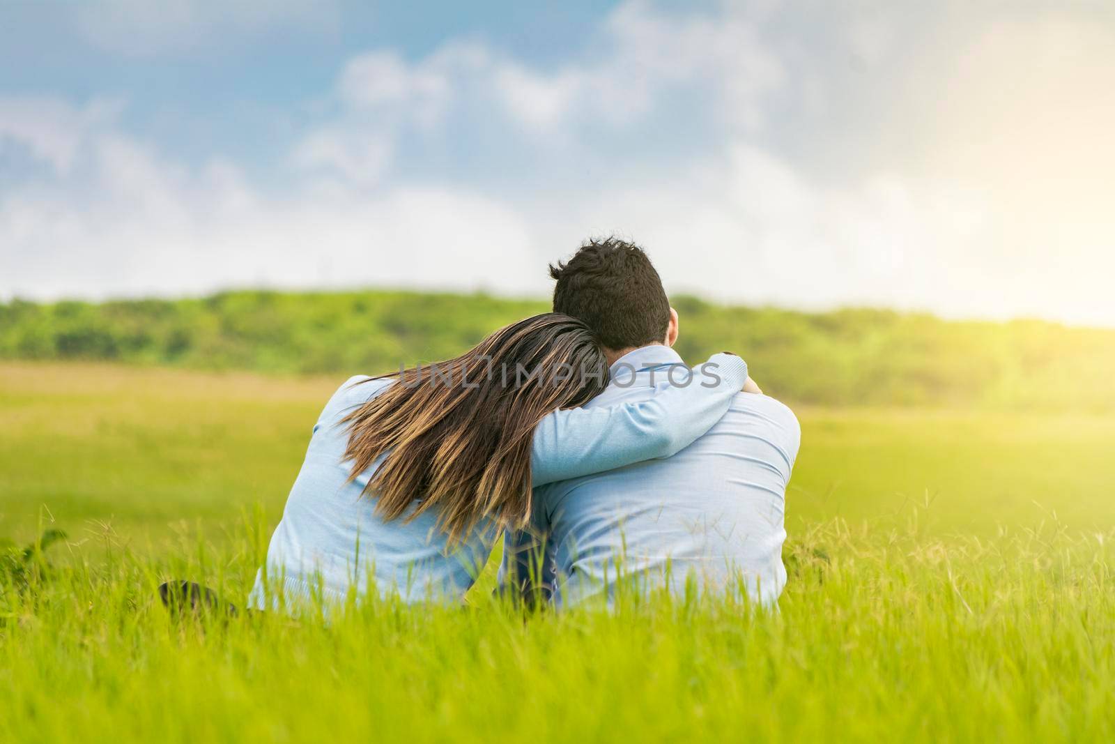 Romantic couple sitting on the grass hugging from the back, rear view of a couple in love hugging on the grass, A couple in love sitting on the grass hugging from the back