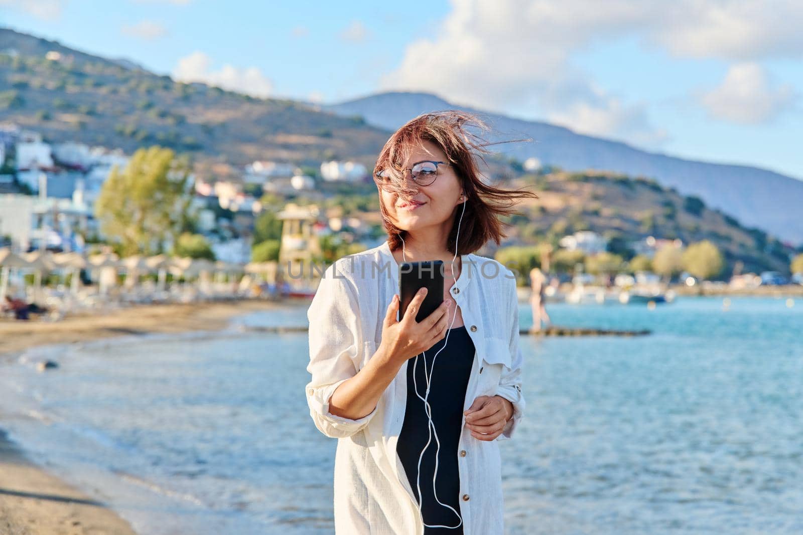 Middle-aged smiling woman relaxing on beach wearing headphones with smartphone. Mature female listening audio podcast, watching video. Leisure technology summer vacations, seaside resorts, people 40s