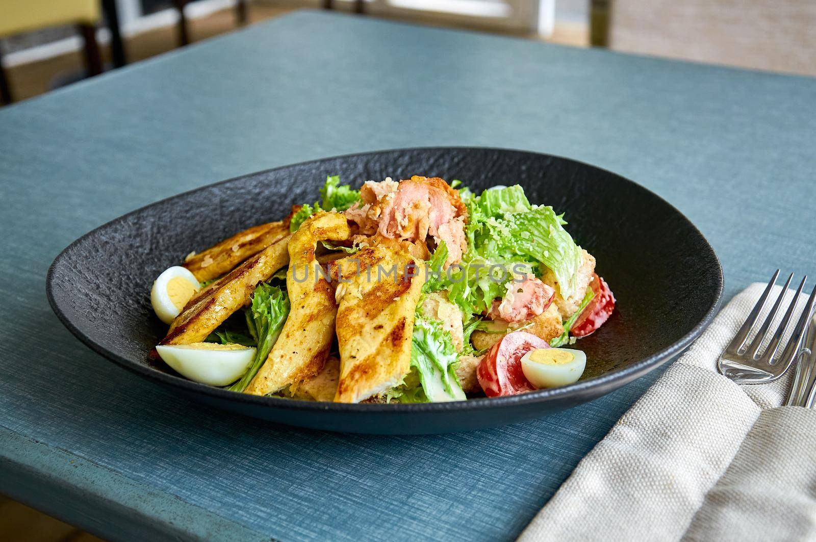 Fresh Caesar salad with chicken in a black bowl on the veranda of the restaurant.