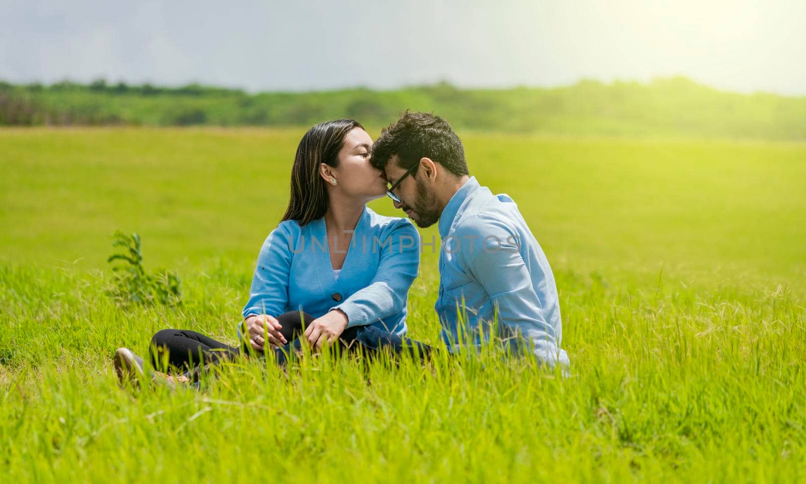 Romantic couple sitting on the grass kissing their foreheads, A pretty girl kissing her boyfriend's forehead in the field, A couple in love sitting on the field kissing their foreheads by isaiphoto