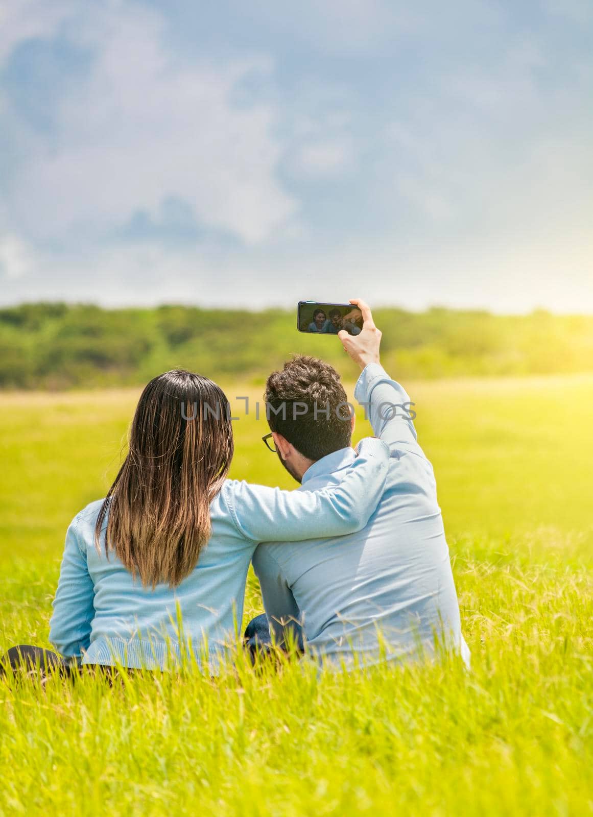 People in love taking selfies in the field with their smartphone, Smiling couple in love sitting on the grass taking selfies, Young couple in love taking a selfie in the field