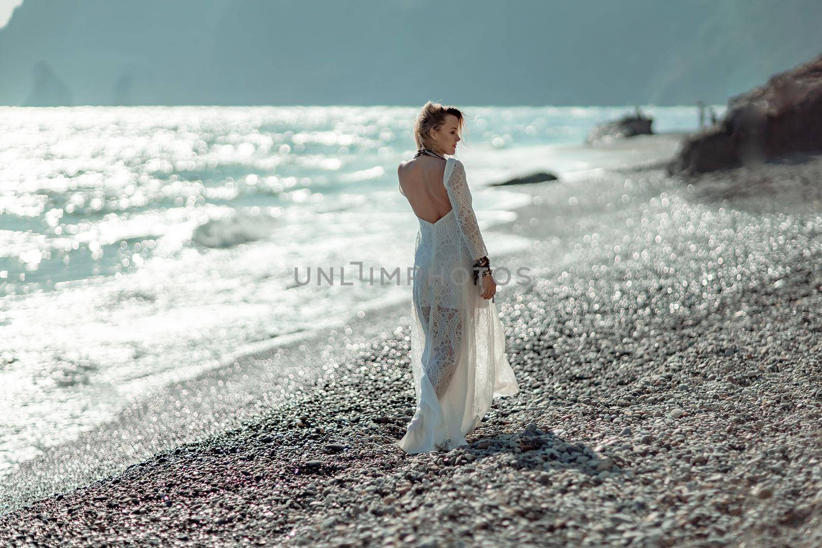 Middle aged woman looks good with blond hair, boho style in white long dress on the beach decorations on her neck and arms. by Matiunina