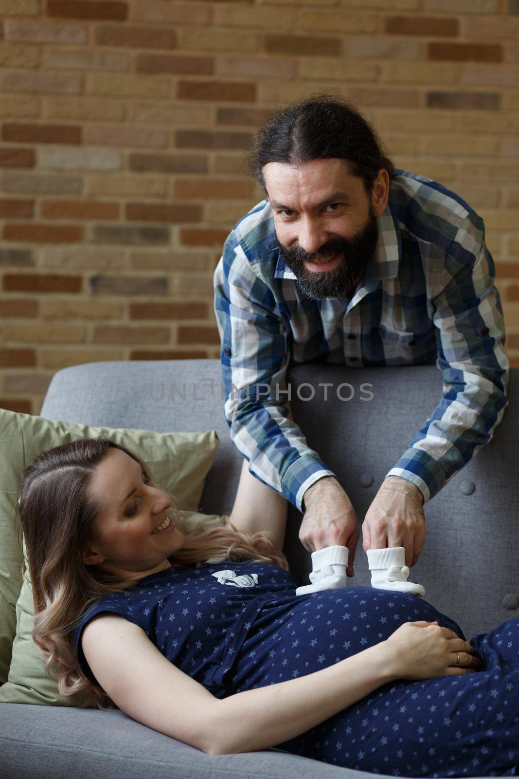 A man and his pregnant wife pose on a sofa in their home. by BY-_-BY