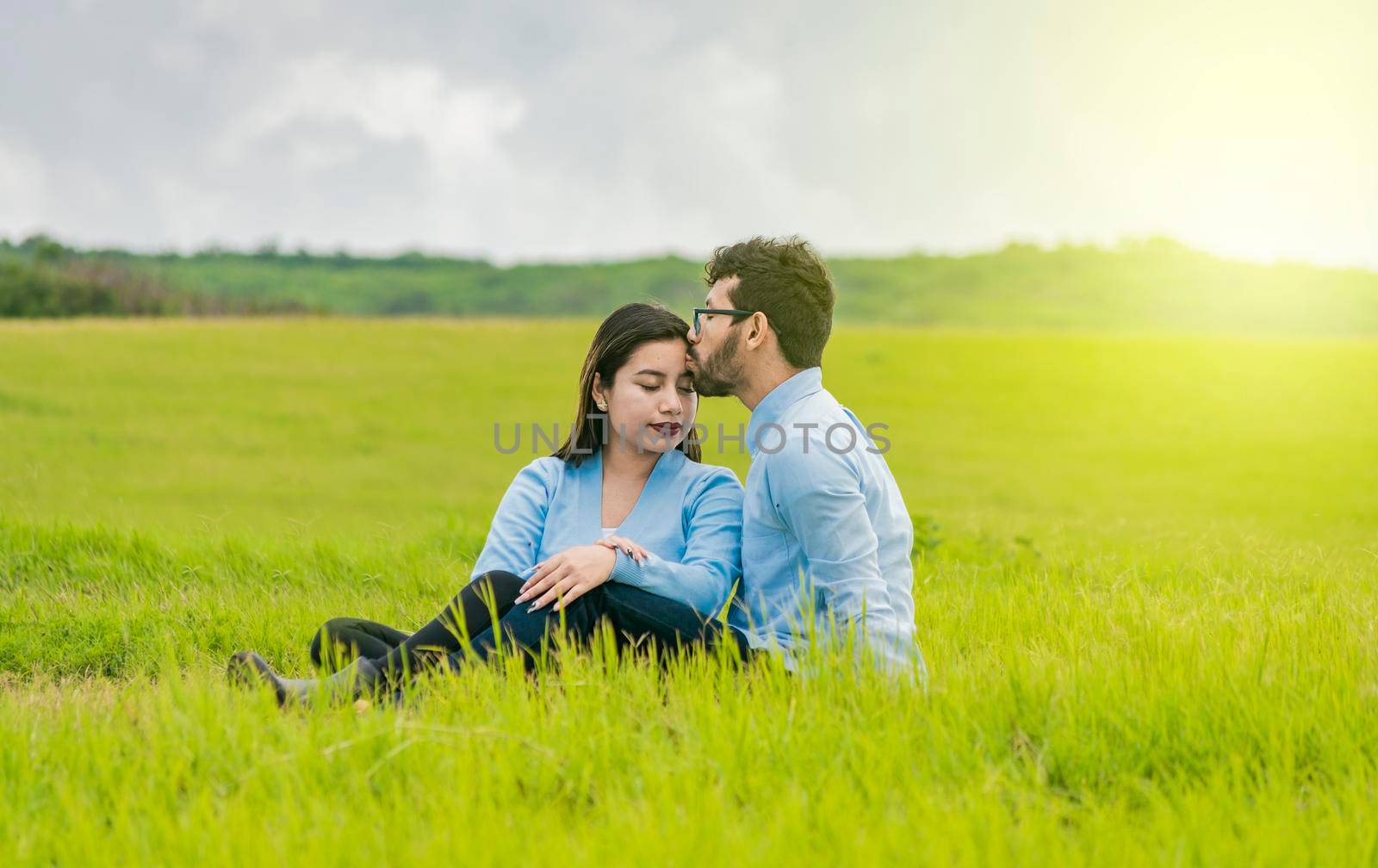 A couple in love sitting in the field kissing their foreheads, A man kissing his girlfriend's forehead in the field, Romantic couple sitting in the grass kissing their foreheads