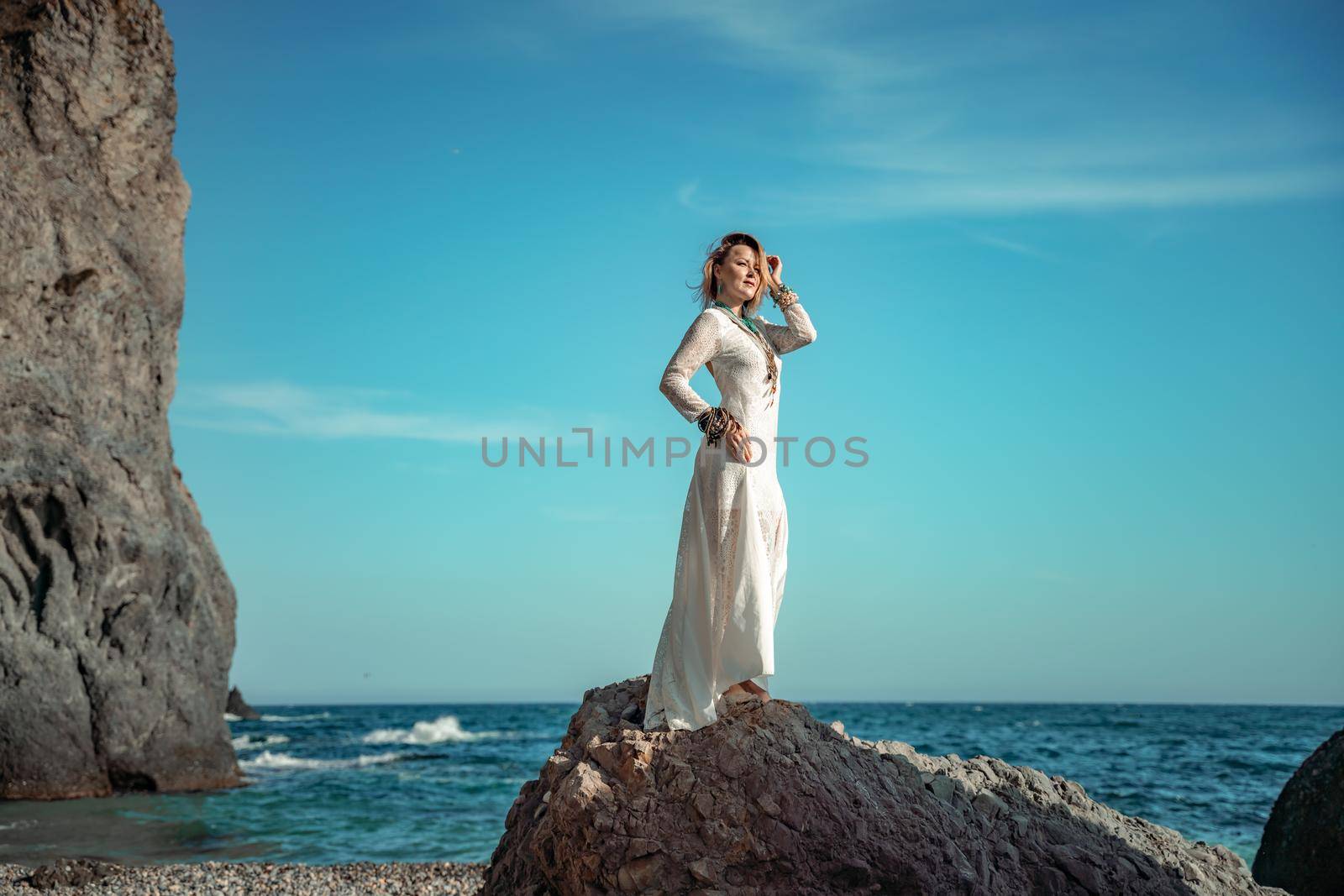 Middle aged woman looks good with blond hair, boho style in white long dress on the beach decorations on her neck and arms