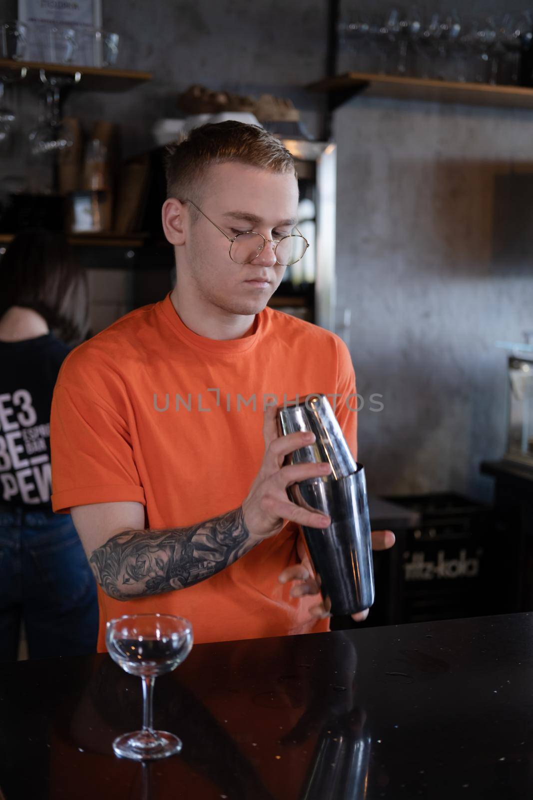 stylish young man hipster in orange t-shirt making mixing a cocktail in a dark loft cafe. alcohol drink in modern bar.