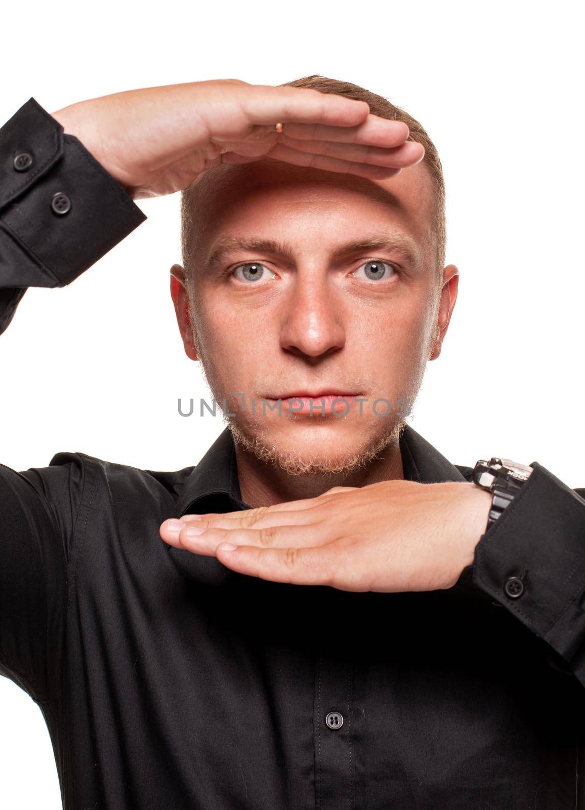 Handsome young blond man in a black shirt is making faces and looking at the camera through his hands, isolated on a white background