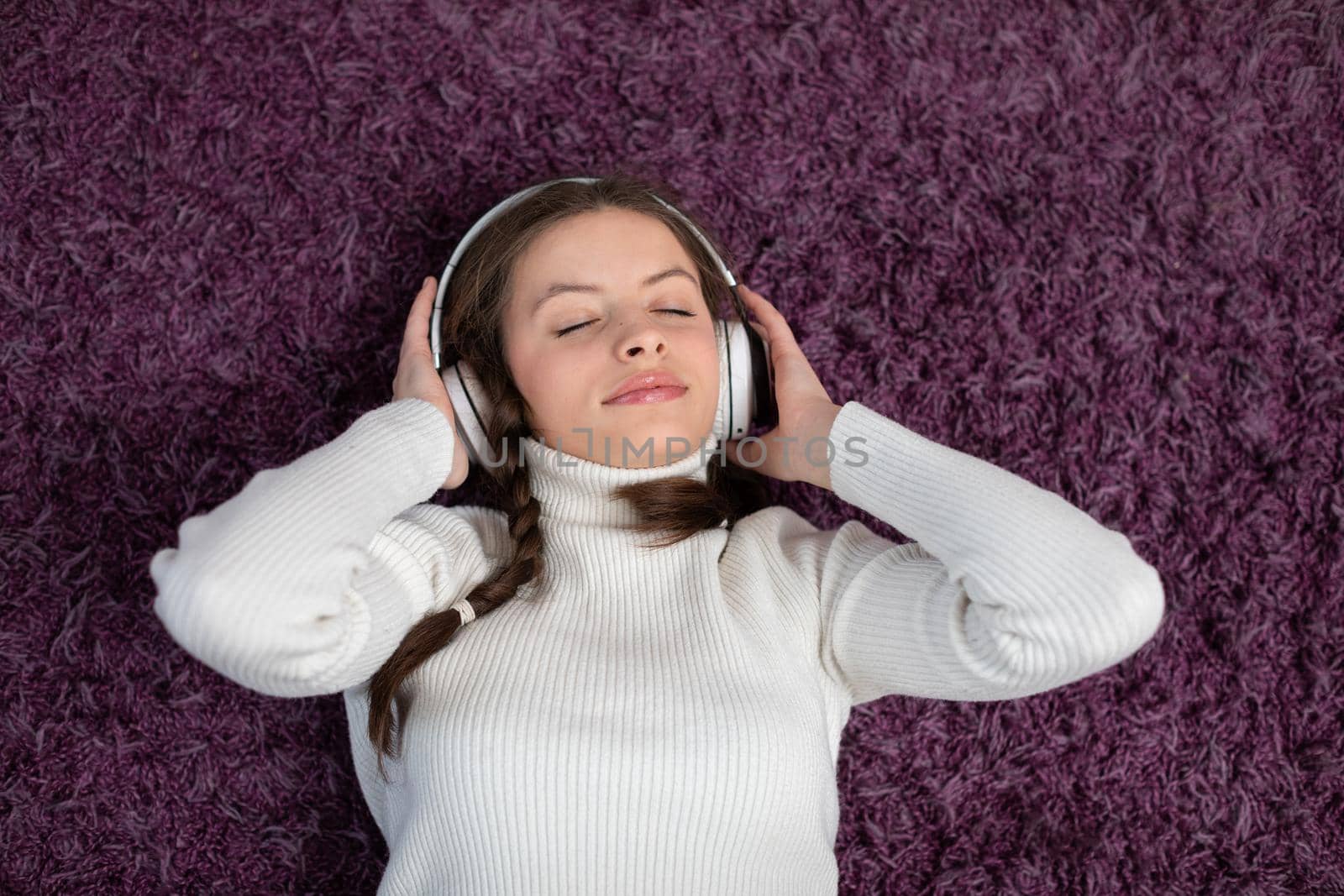 Teenage girl in braids lies on purple carpet with wireless headphones and listens to music. A gentle girlish smile.