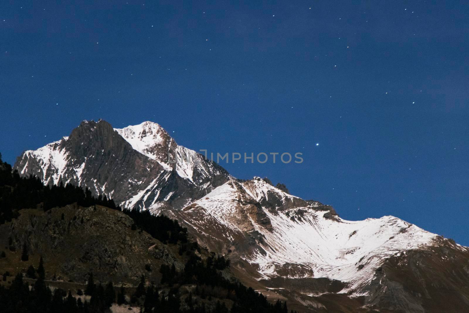 A rocky mountain top with a dusting of snow is illuminated by a full moon out of shot, as stars twinkle above in the midnight blue sky