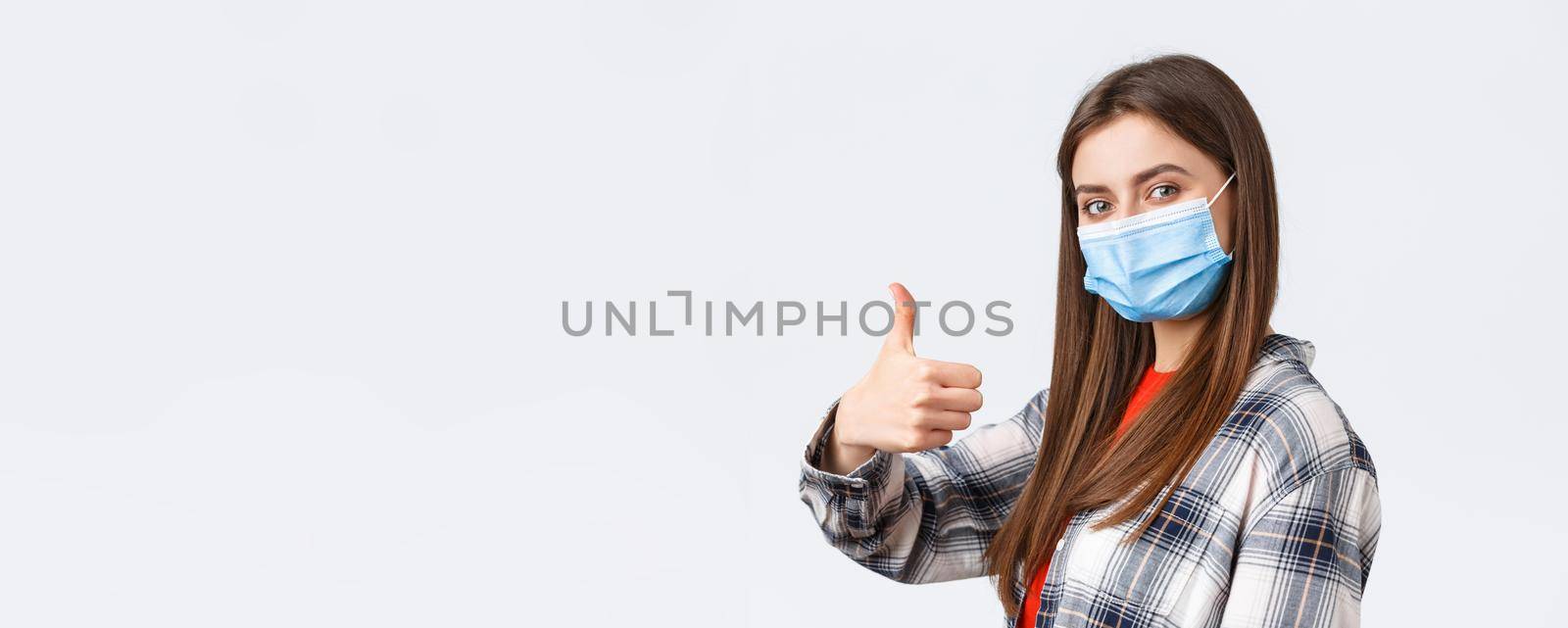 Coronavirus outbreak, leisure on quarantine, social distancing and emotions concept. Cheerful young woman in medical mask stand in profile, turn to camera and thumb-up in approval.
