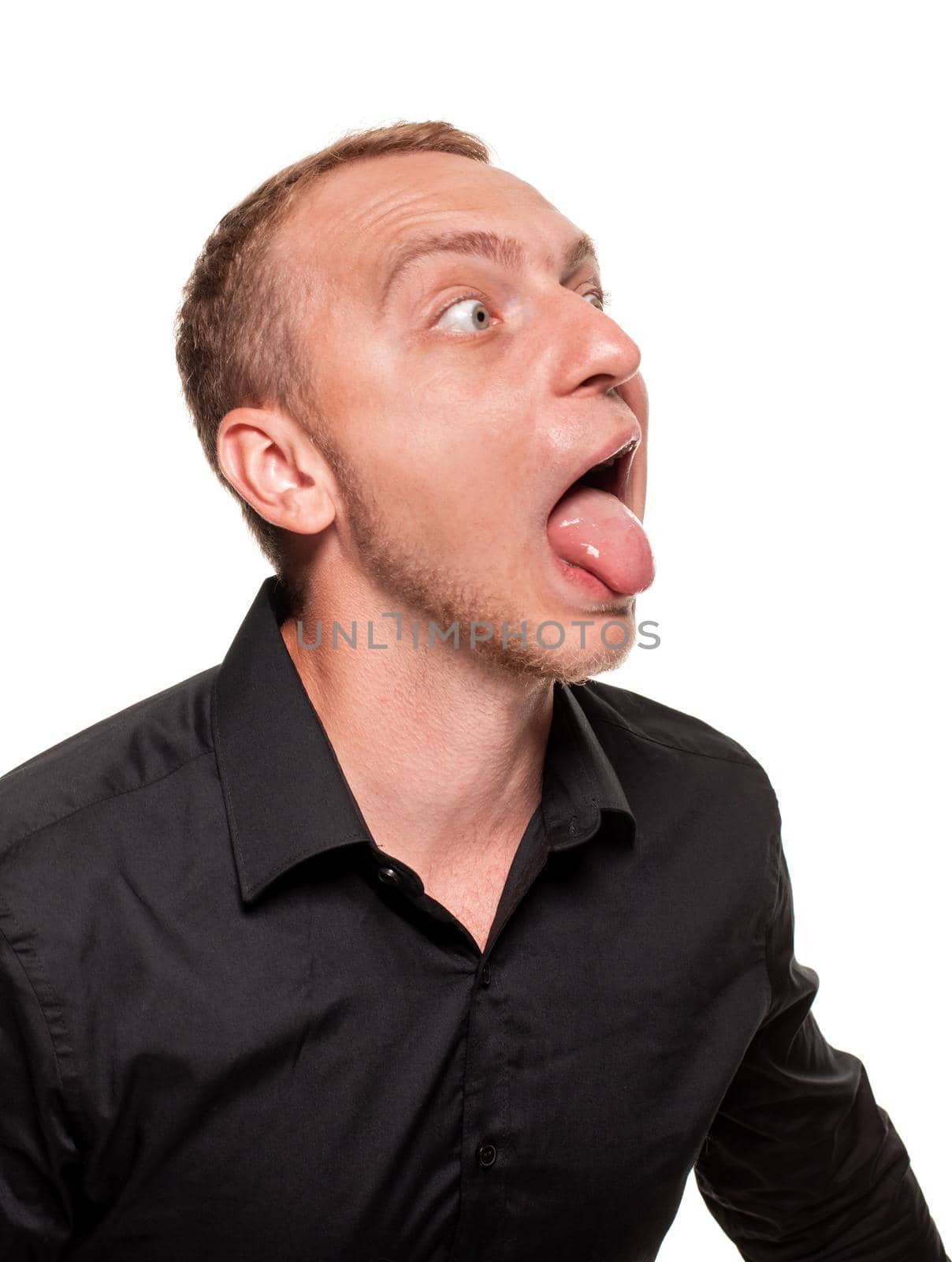 Handsome young blond man in a black shirt is making faces, showing his tongue and looking away, isolated on a white background