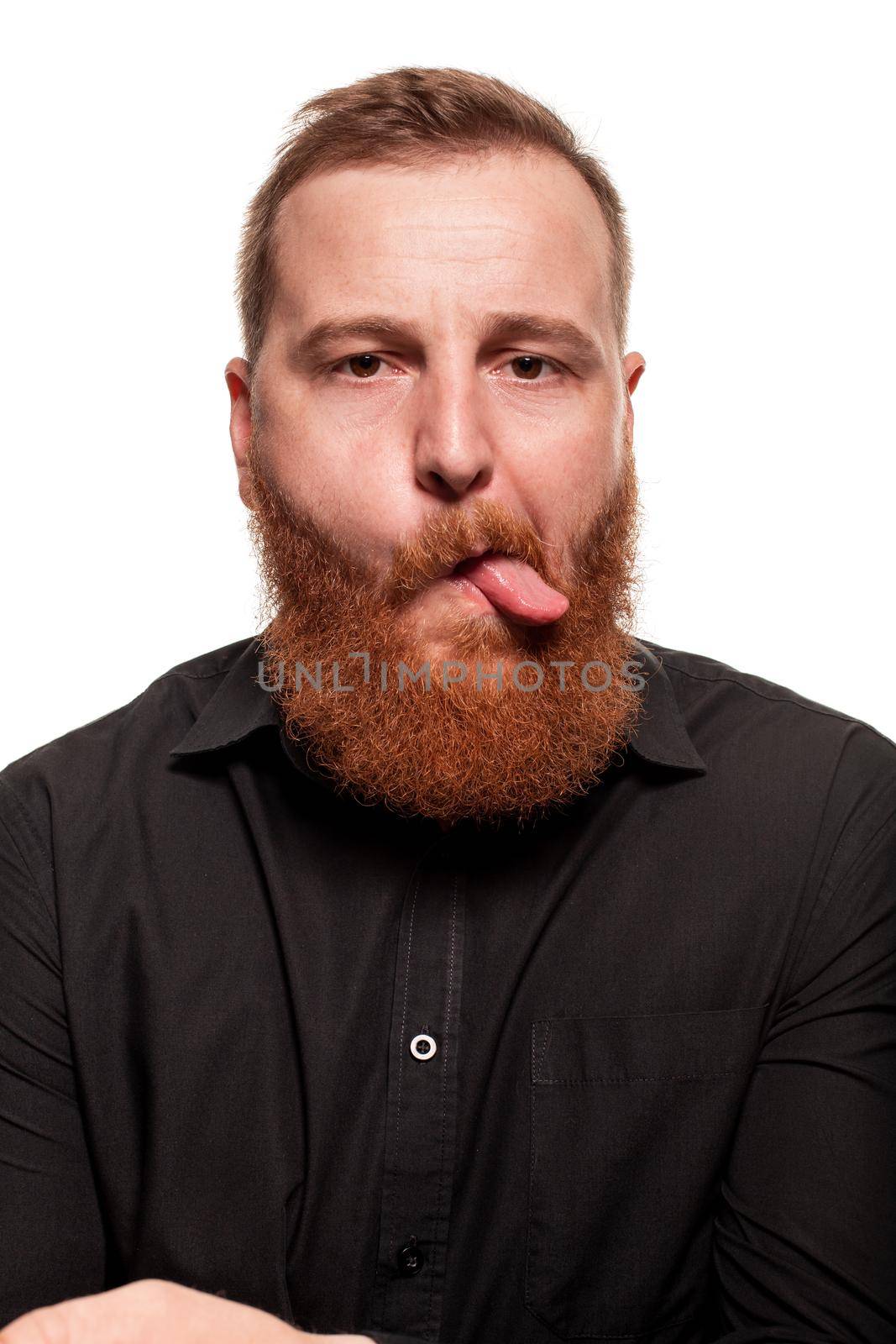 Portrait of a young, redheaded man with a beard in a black shirt, showing his tongue at the camera, isolated on a white background