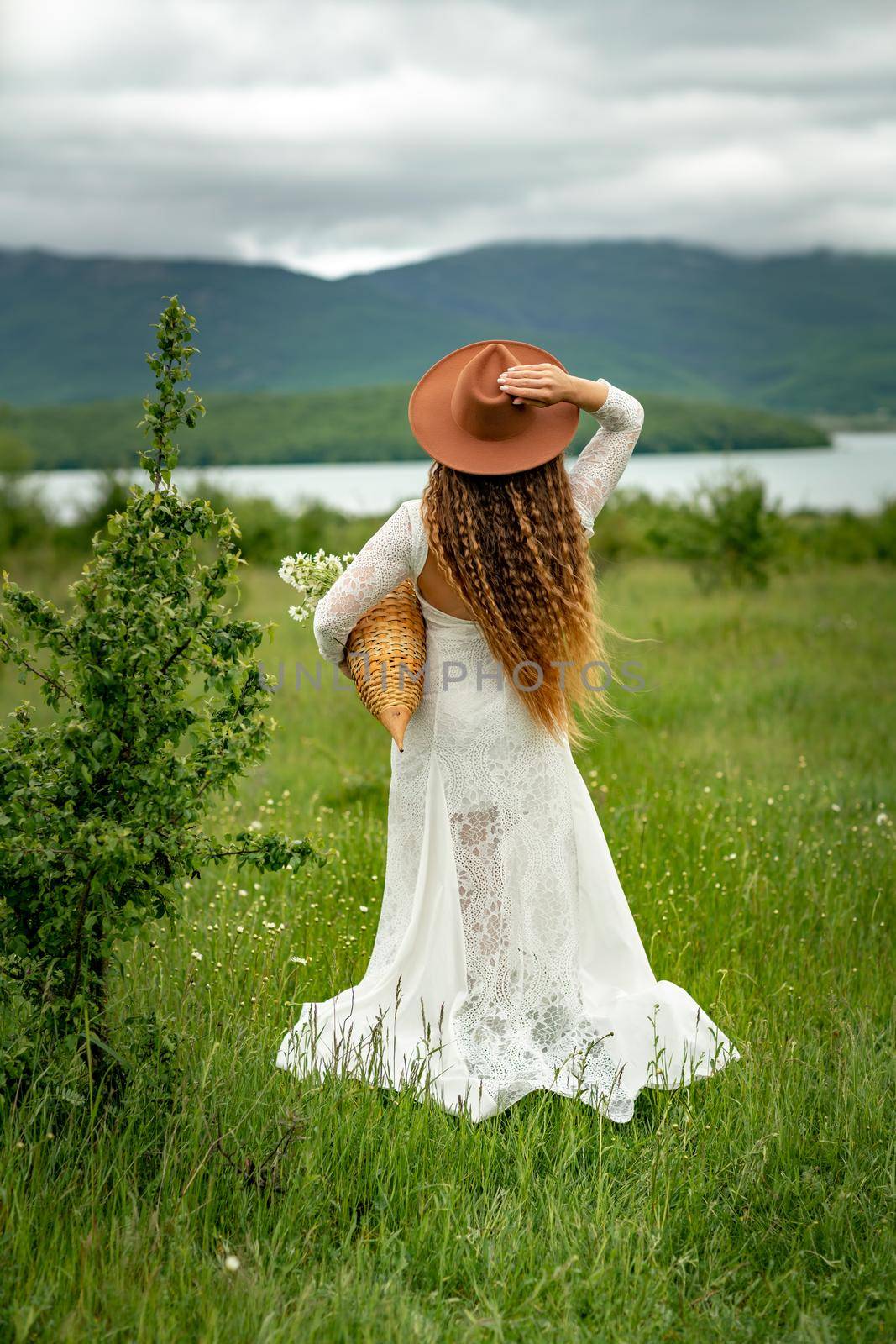 A middle-aged woman in a white dress and brown hat stands with her back on a green field and holds a basket in her hands with a large bouquet of daisies. In the background there are mountains and a lake. by Matiunina