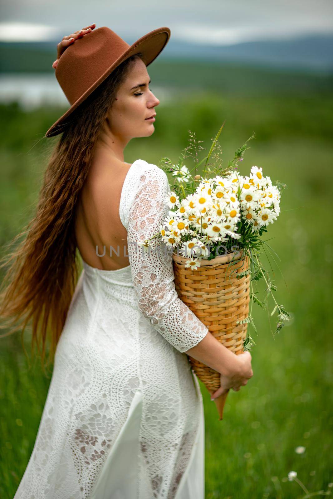 A middle-aged woman in a white dress and brown hat stands on a green field and holds a basket in her hands with a large bouquet of daisies. In the background there are mountains and a lake. by Matiunina
