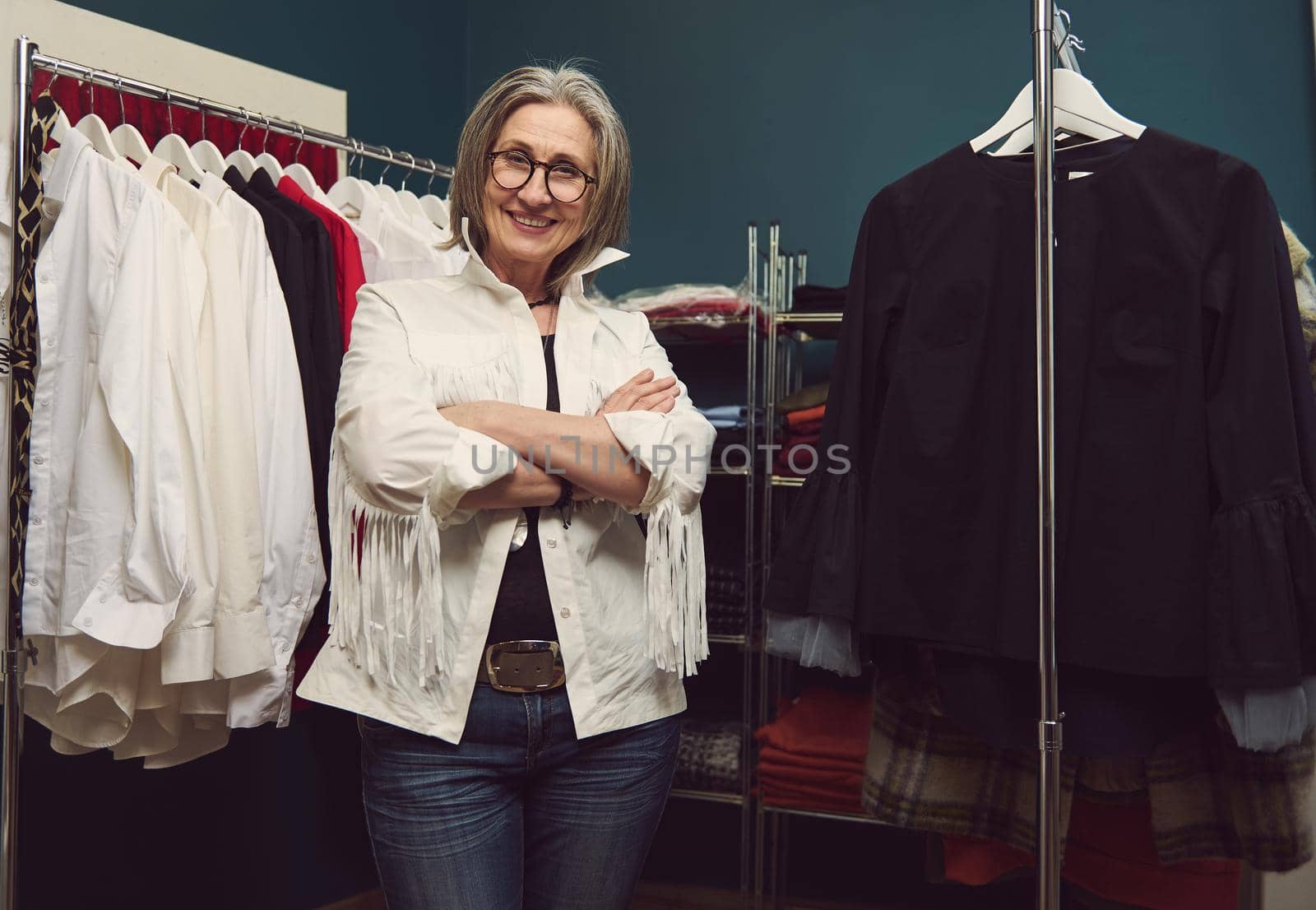 Confident successful elderly European woman seamstress, dressmaker, tailor posing in own fashion design atelier. Clothing designer between hangers with clothing garment in tailoring workshop