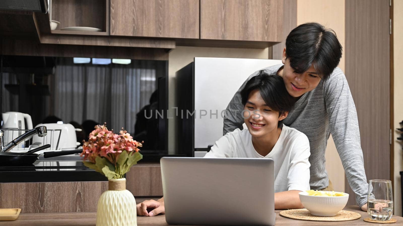 Adorable gay couple spending time, surfing internet on laptop together at kitchen table. LGBT, pride, relationships and equality concept by prathanchorruangsak