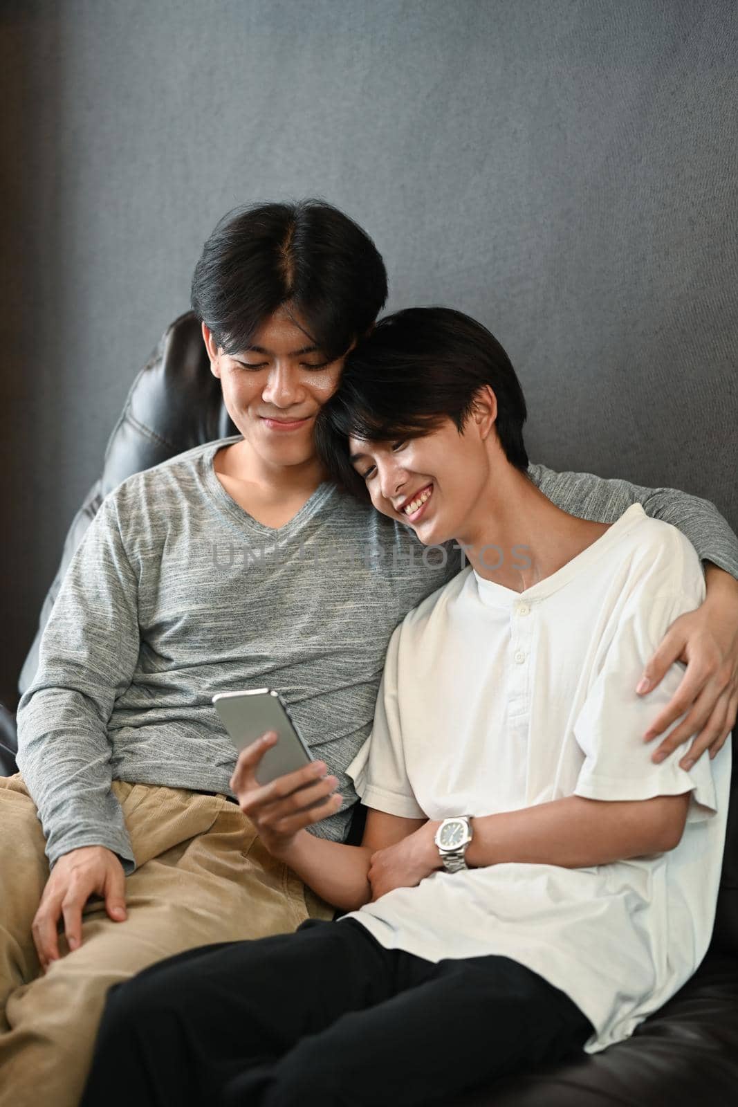 Happy same sex male couple using smart phone on sofa, spending time at home. LGBT, pride, relationships and equality concept by prathanchorruangsak