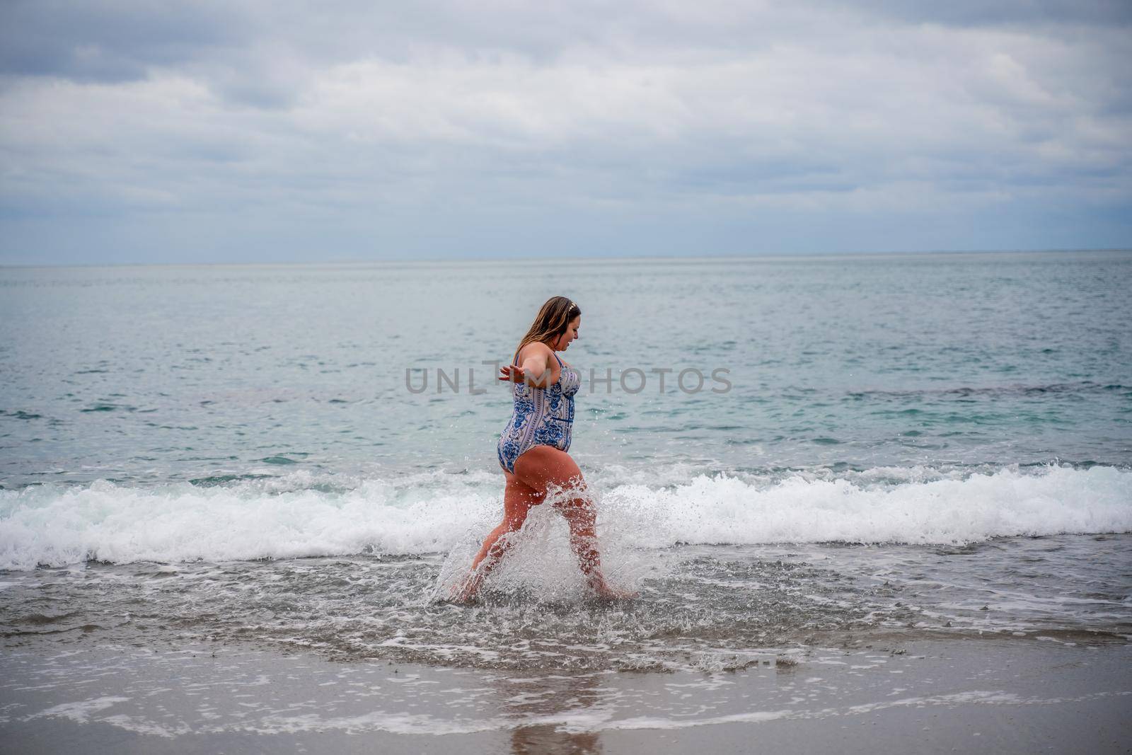 A plump woman in a bathing suit enters the water during the surf. Alone on the beach, Gray sky in the clouds, swimming in winter