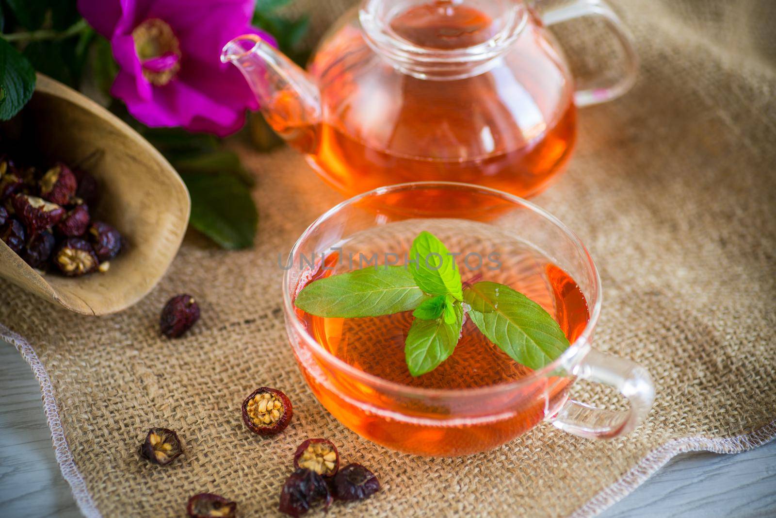 brewed rosehip tea in a glass teapot with rosehip flowers and mint, on a wooden table.