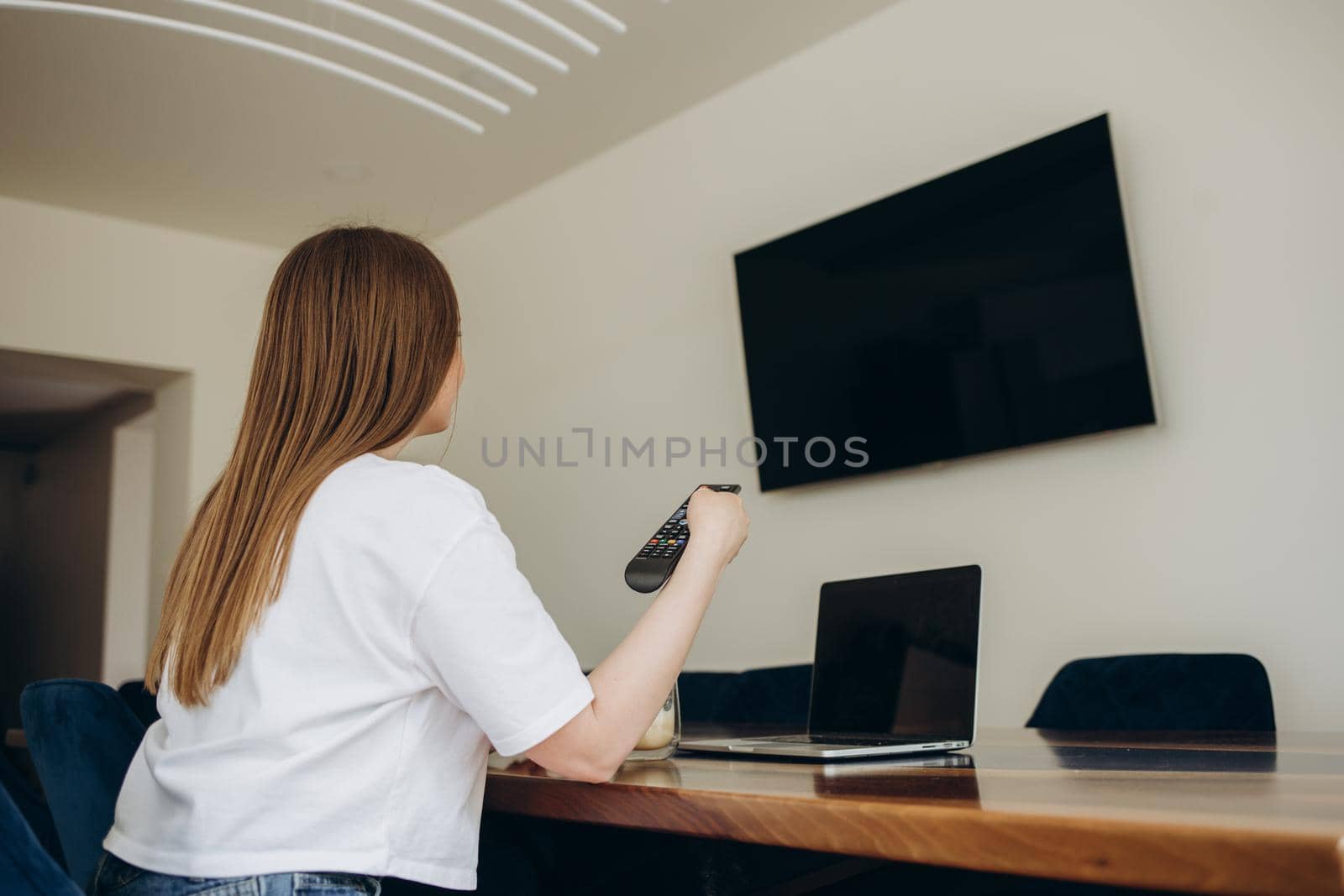 young beautiful woman sitting in the kitchen near the laptop, turns on the TV remote control to the TV