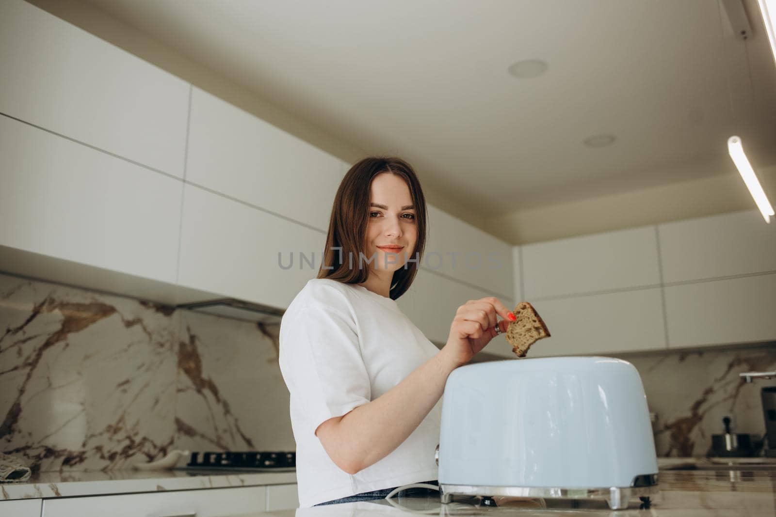 Pretty young woman preparing a toast at home in the kitchen. Modern interior, furniture for the kitchen. Cook at home. Healthy eating and diet. by fentonroma